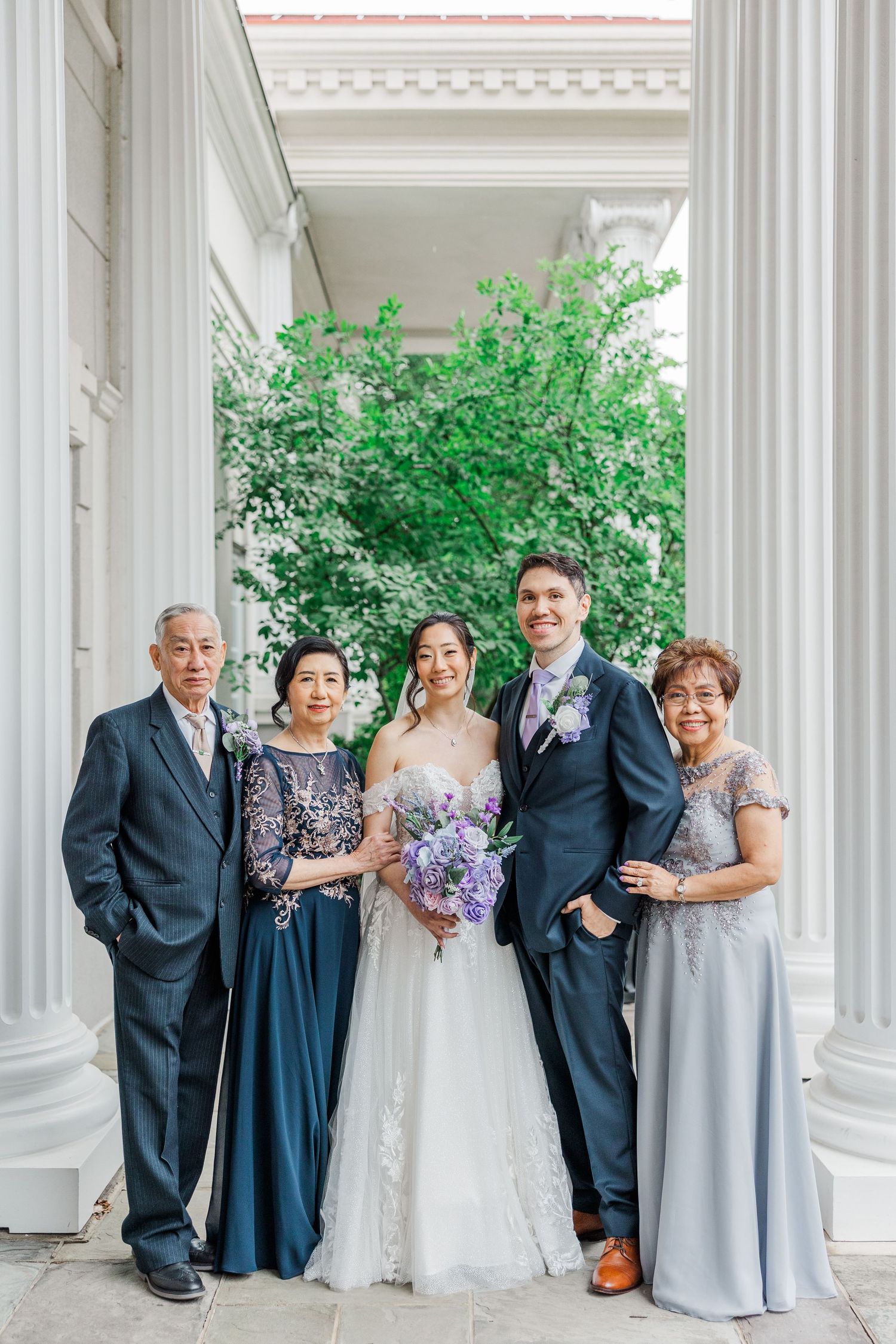 Bride and groom pose with family between white columns, surrounded by lush greenery, bride holding purple bouquet.
