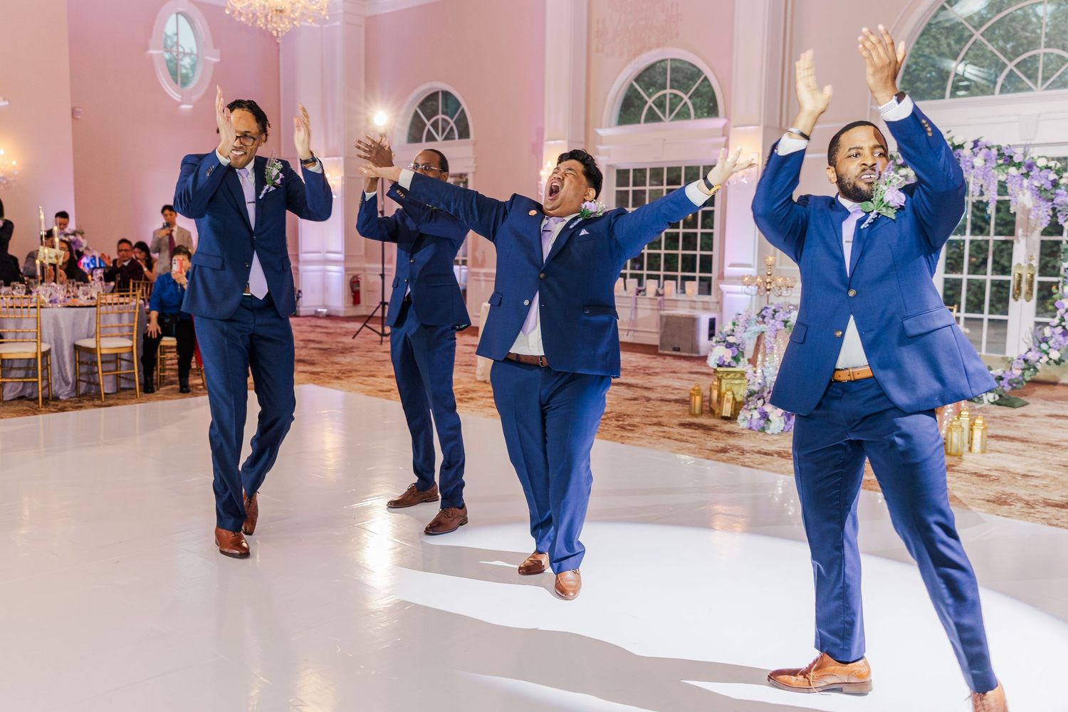 Four groomsmen in navy blue suits performing a choreographed dance on a white ballroom floor at a wedding reception.
