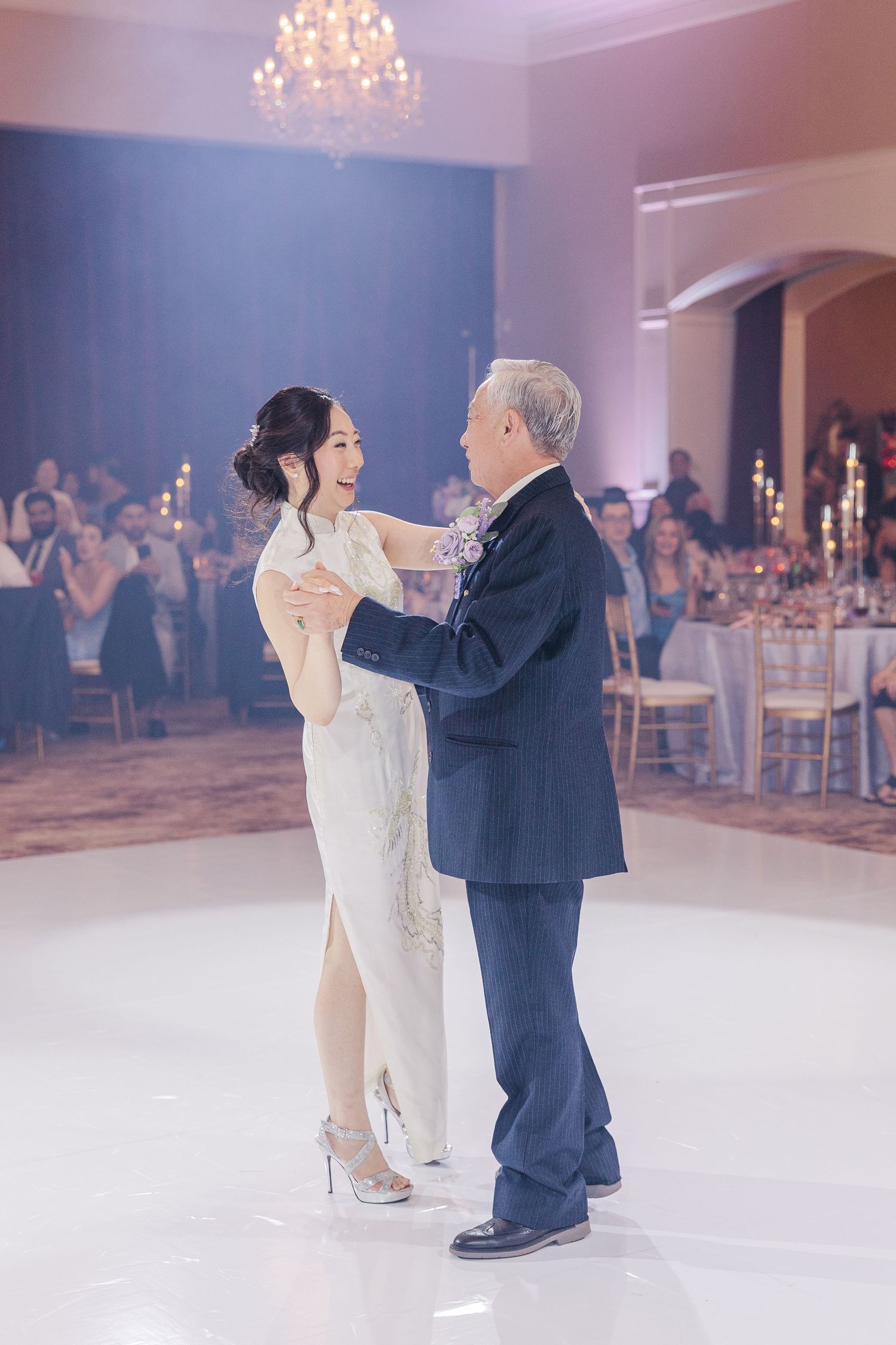 Bride in white qipao shares father-daughter dance at elegant wedding reception with chandelier and candlelit tables.