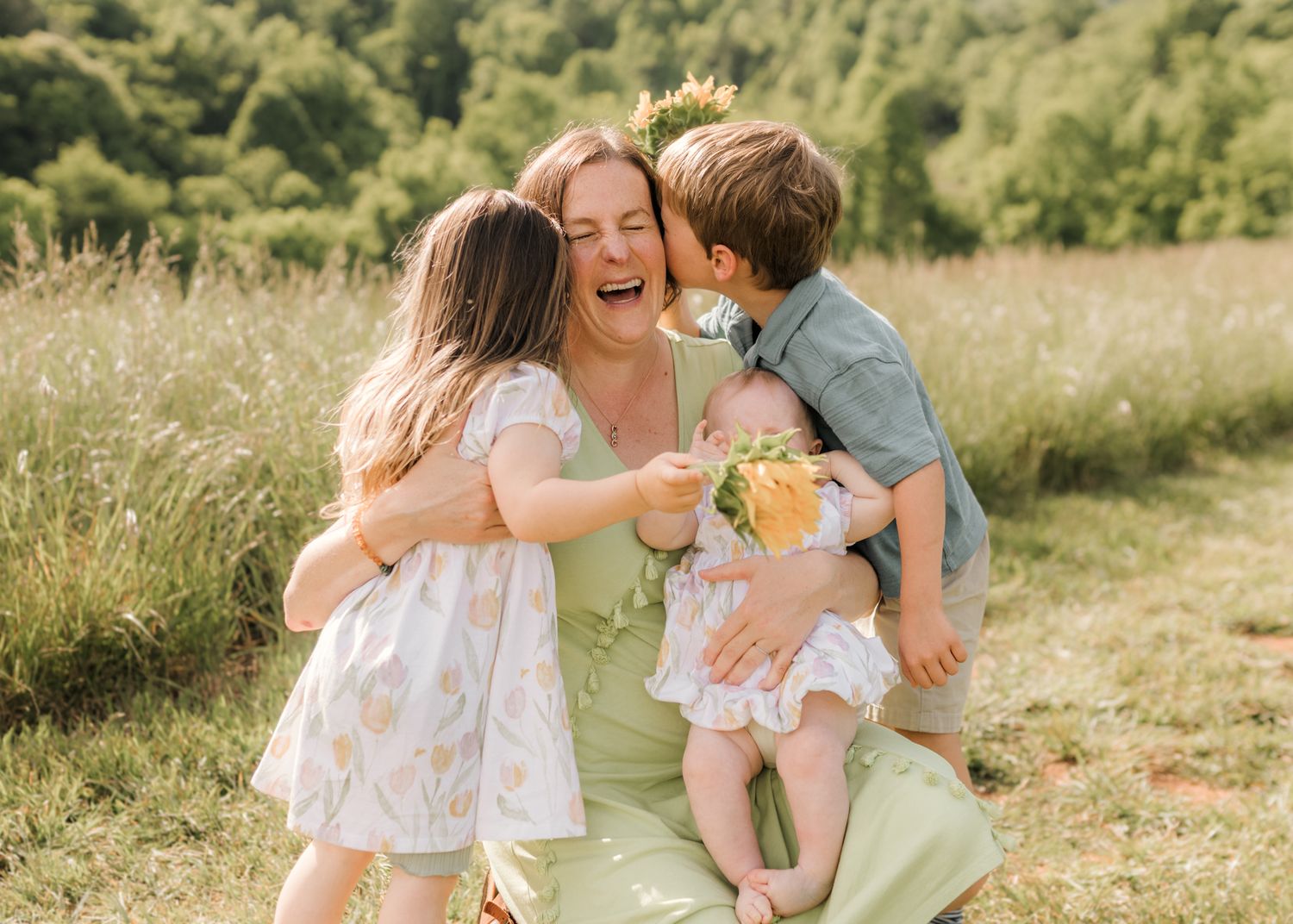 A mother sits in a sunny meadow smiling as her two young children hug and kiss her cheeks outdoors.