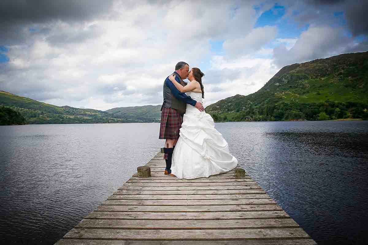 Bride and groom kissing on a wooden pier beside lake Ullswater with the groom wearing traditional tartan kilt, scenic hills behind.