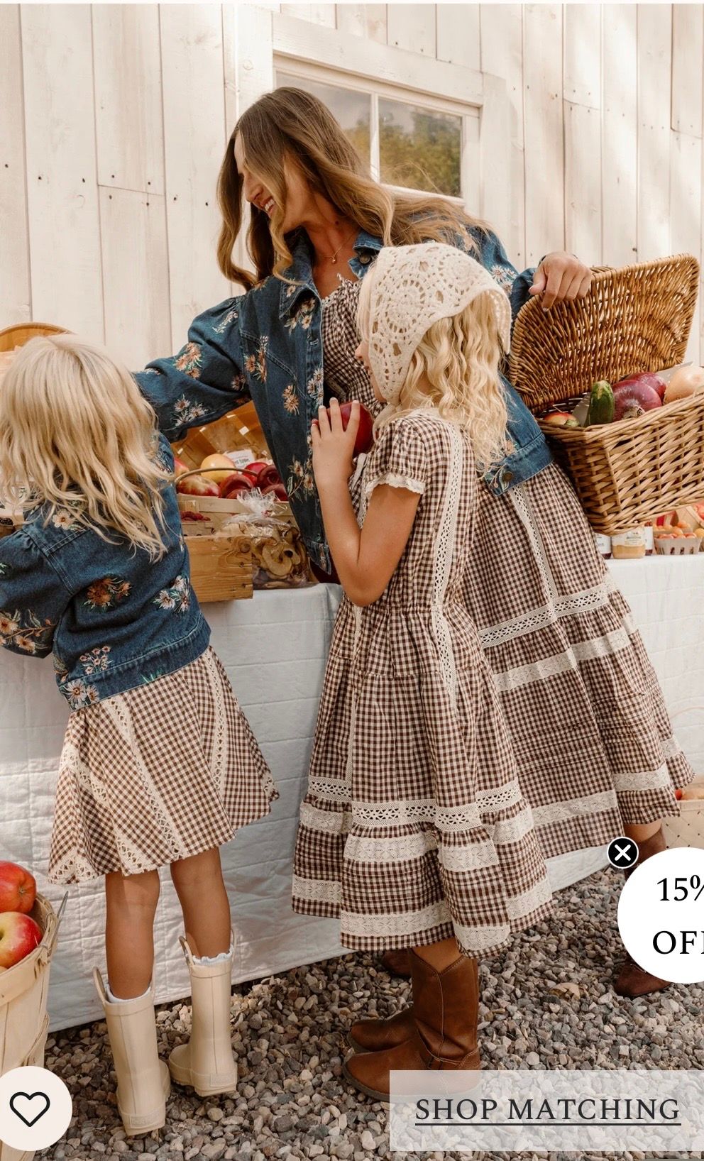 Mother and two girls in matching brown gingham dresses shop at a rustic market stall with wicker baskets.