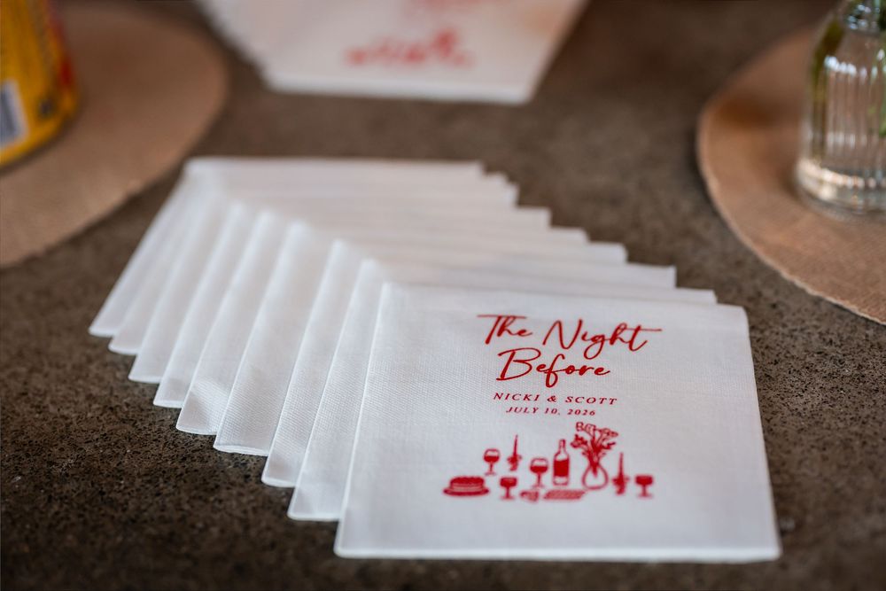 Custom white cocktail napkins printed in red with "The Night Before" wedding rehearsal dinner design stacked on a bar.
