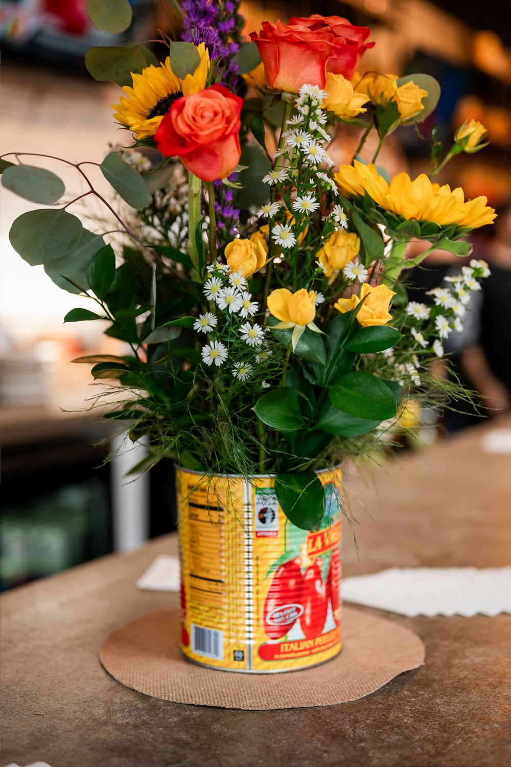Colorful flower arrangement with roses, sunflowers, and daisies displayed in a decorative tin can on a wooden surface.