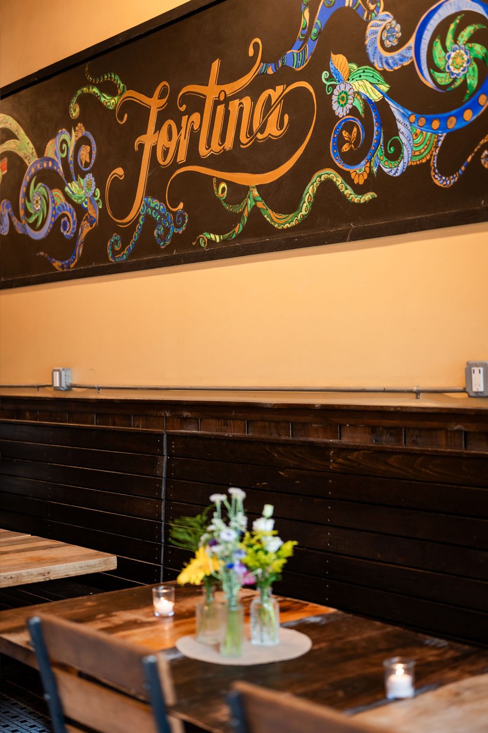 Fortina restaurant interior with decorative chalkboard mural, wooden booths, and rustic tables with fresh flowers.
