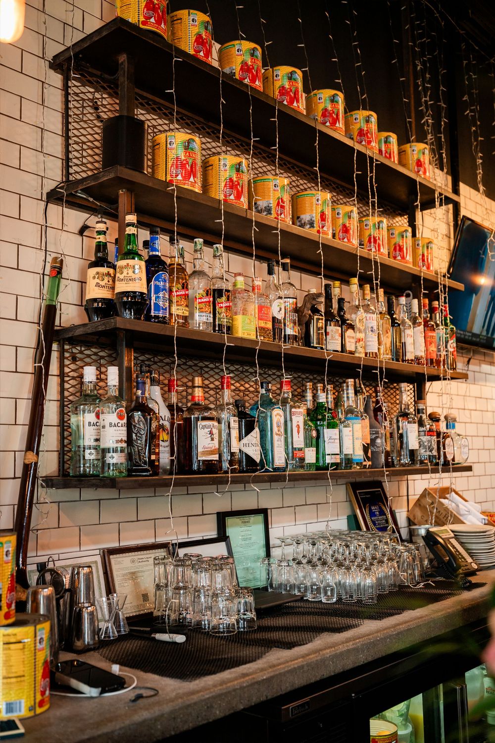 A well-stocked bar shelf displaying various liquor bottles and colorful cans above a counter with clean glassware.
