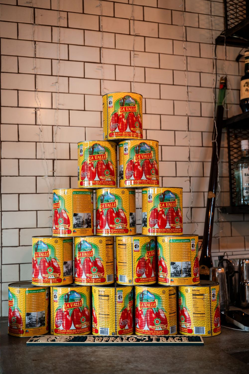 Pyramid stack of La Valle yellow tomato cans displayed on a mat against a white brick wall.