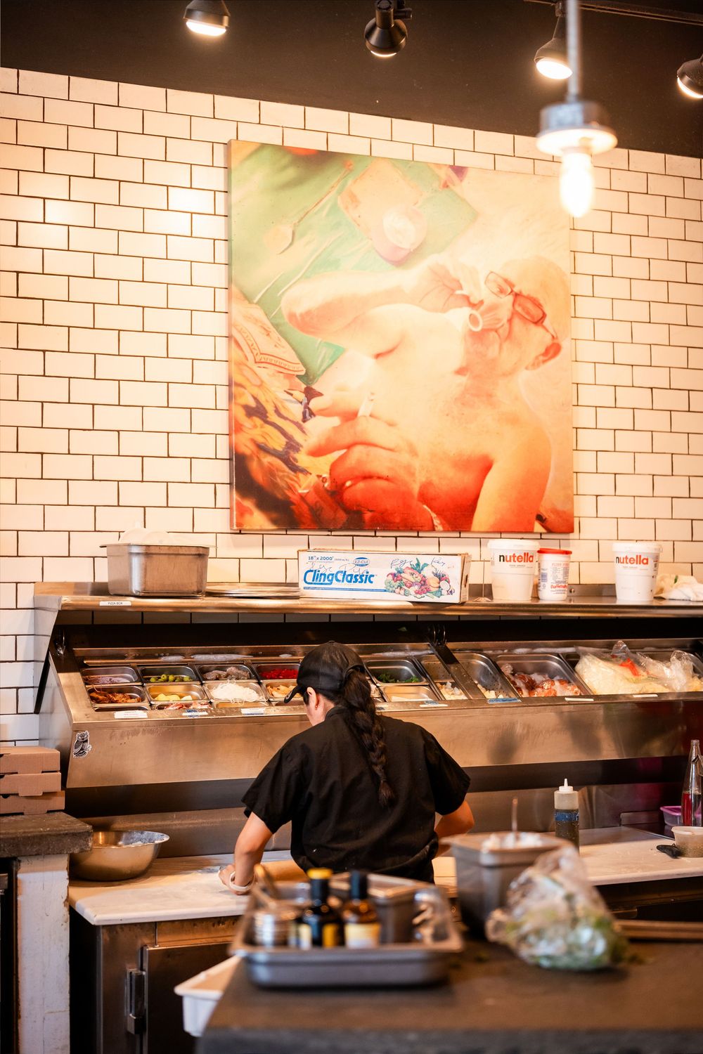 Restaurant kitchen worker in black uniform prepares food at a buffet counter beneath colorful wall art.