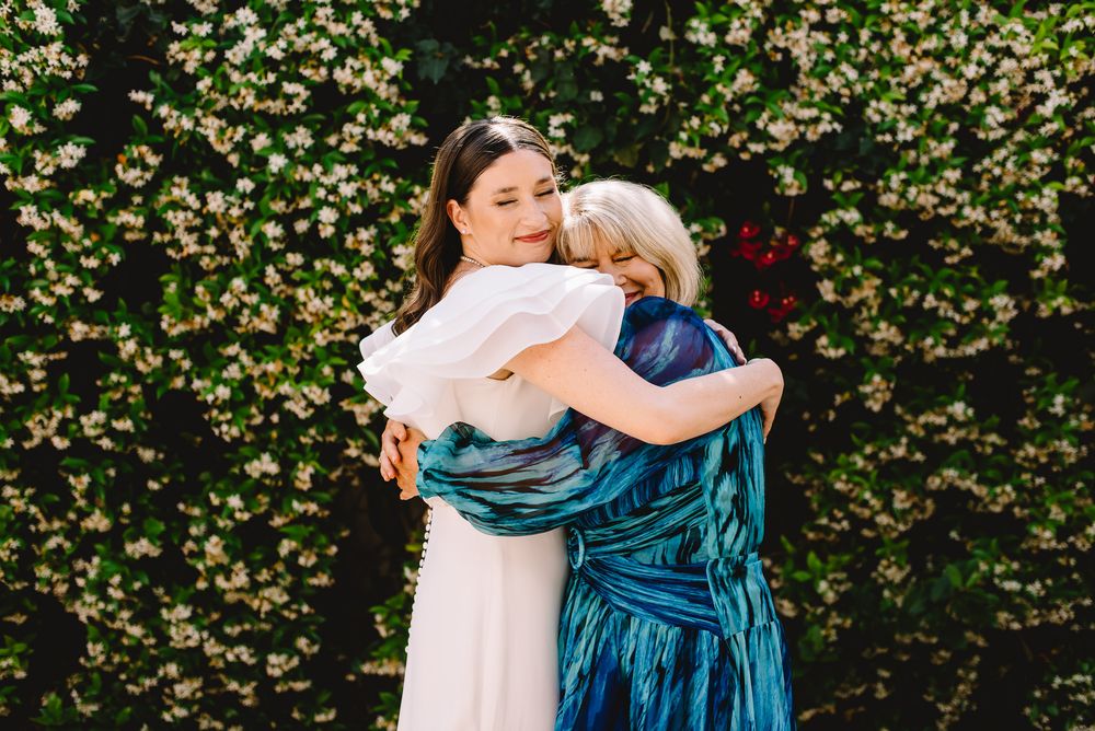 Two women share a warm embrace in front of a lush floral green wall, one wearing a white dress and the other in blue.