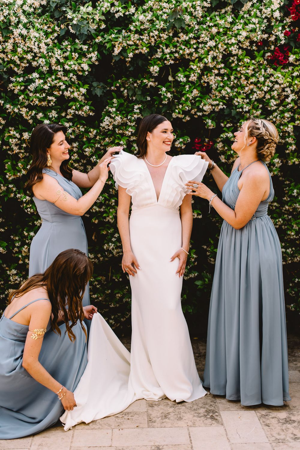 Bride in white ruffle gown assisted by three dusty blue bridesmaids before floral wall backdrop.