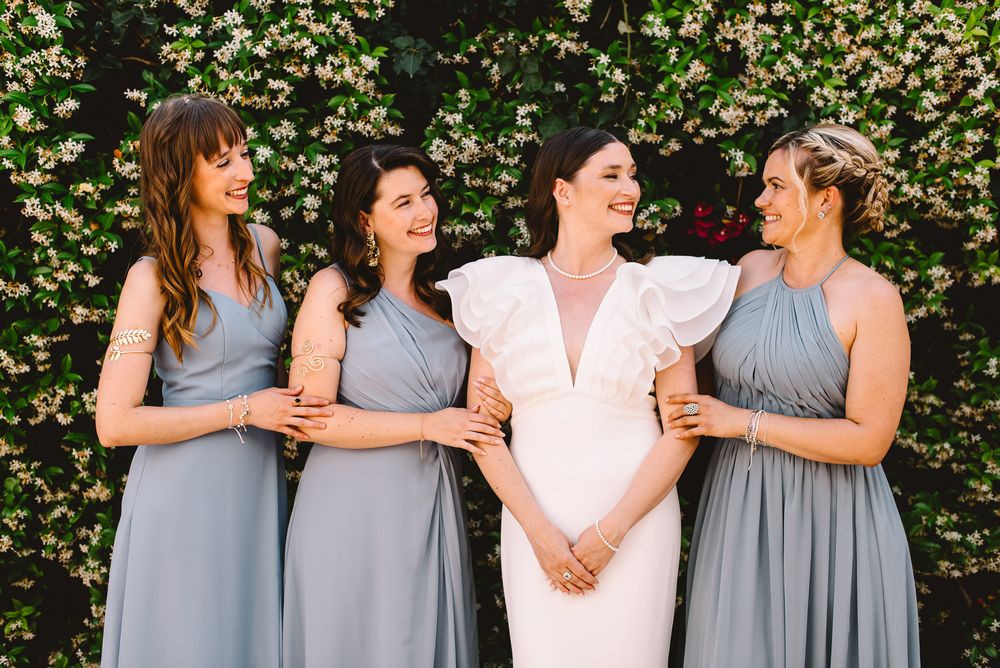 Bride in white ruffled dress with three bridesmaids in sage gray gowns laughing together before floral greenery wall.