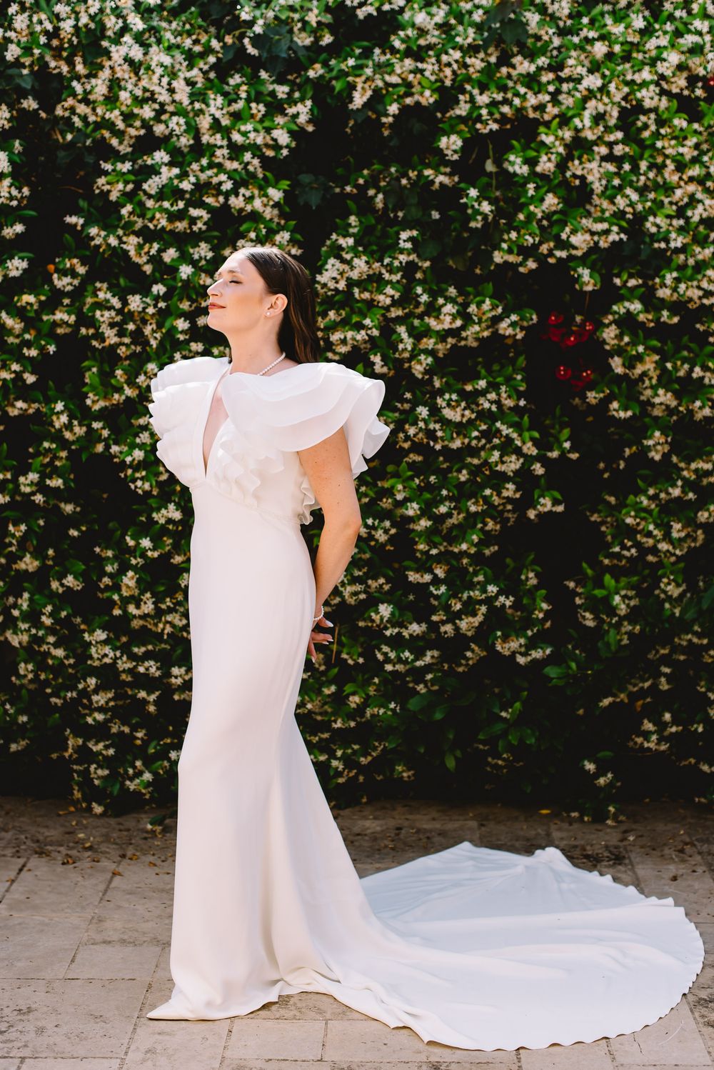 Bride in elegant white ruffle-sleeve gown with train poses before lush flowering hedge backdrop.