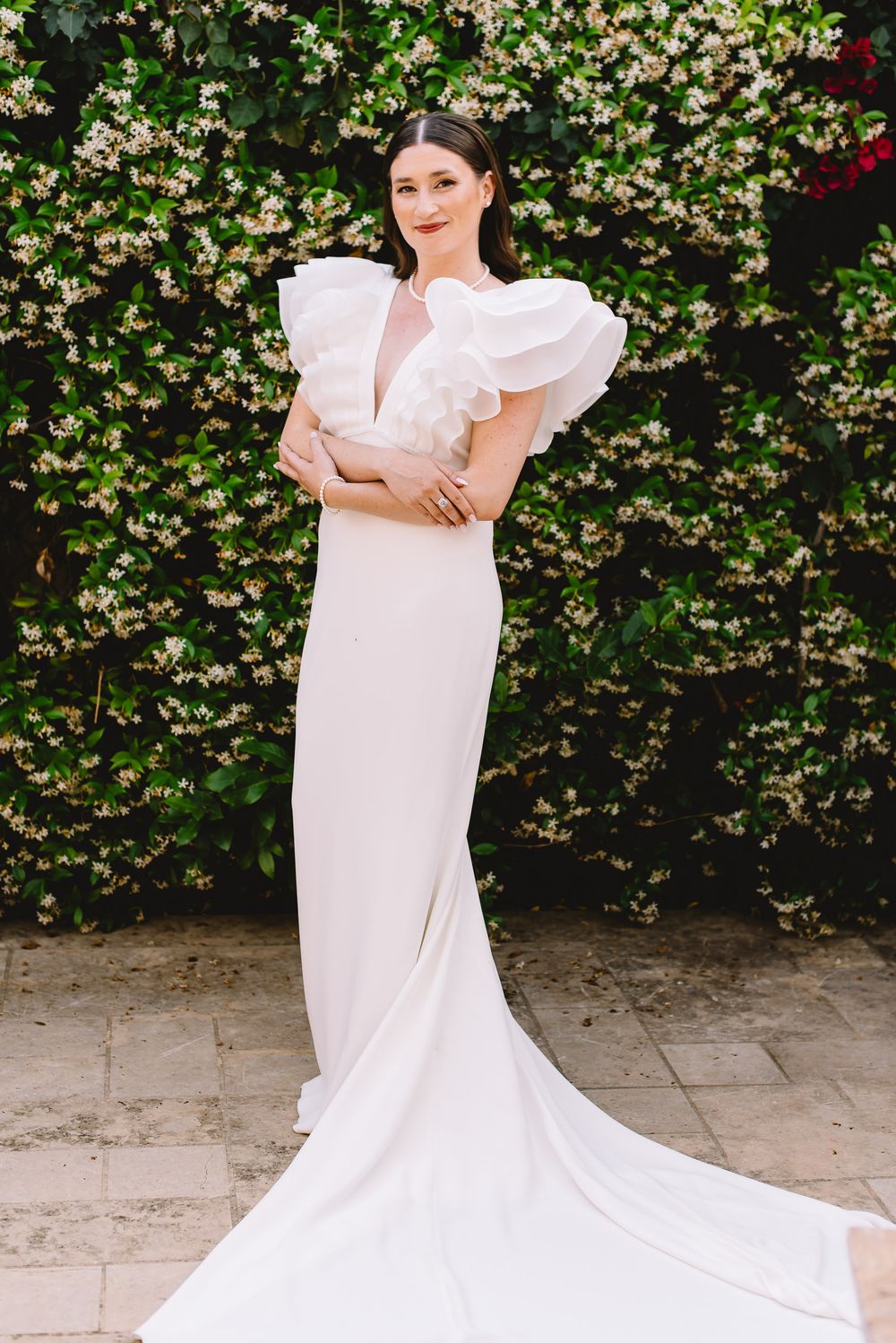 Bride in white gown with ruffled sleeves and train poses before lush green floral wall backdrop.