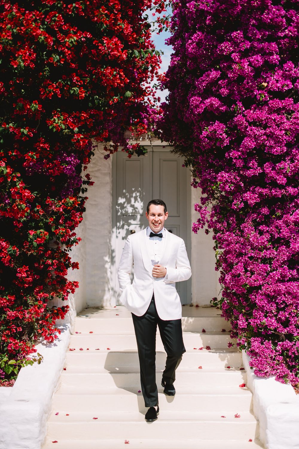 Groom in white tuxedo jacket walks down steps framed by vibrant red and purple bougainvillea flowers.