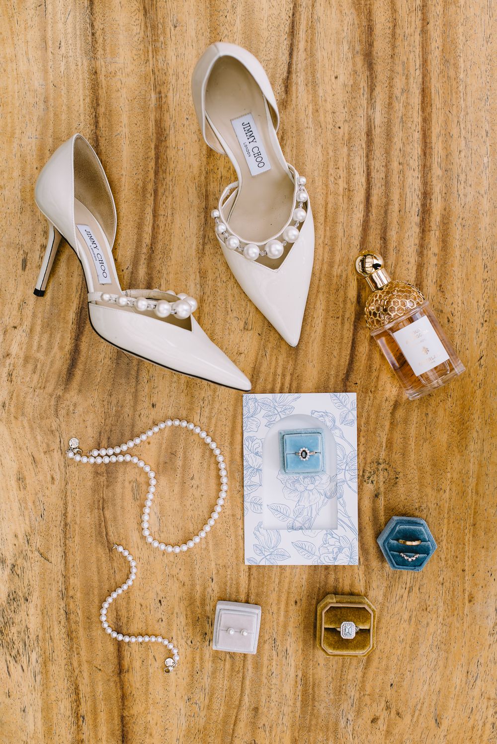 White Jimmy Choo pearl-embellished wedding heels displayed with pearl jewelry, engagement ring, perfume, and blue velvet ring box.