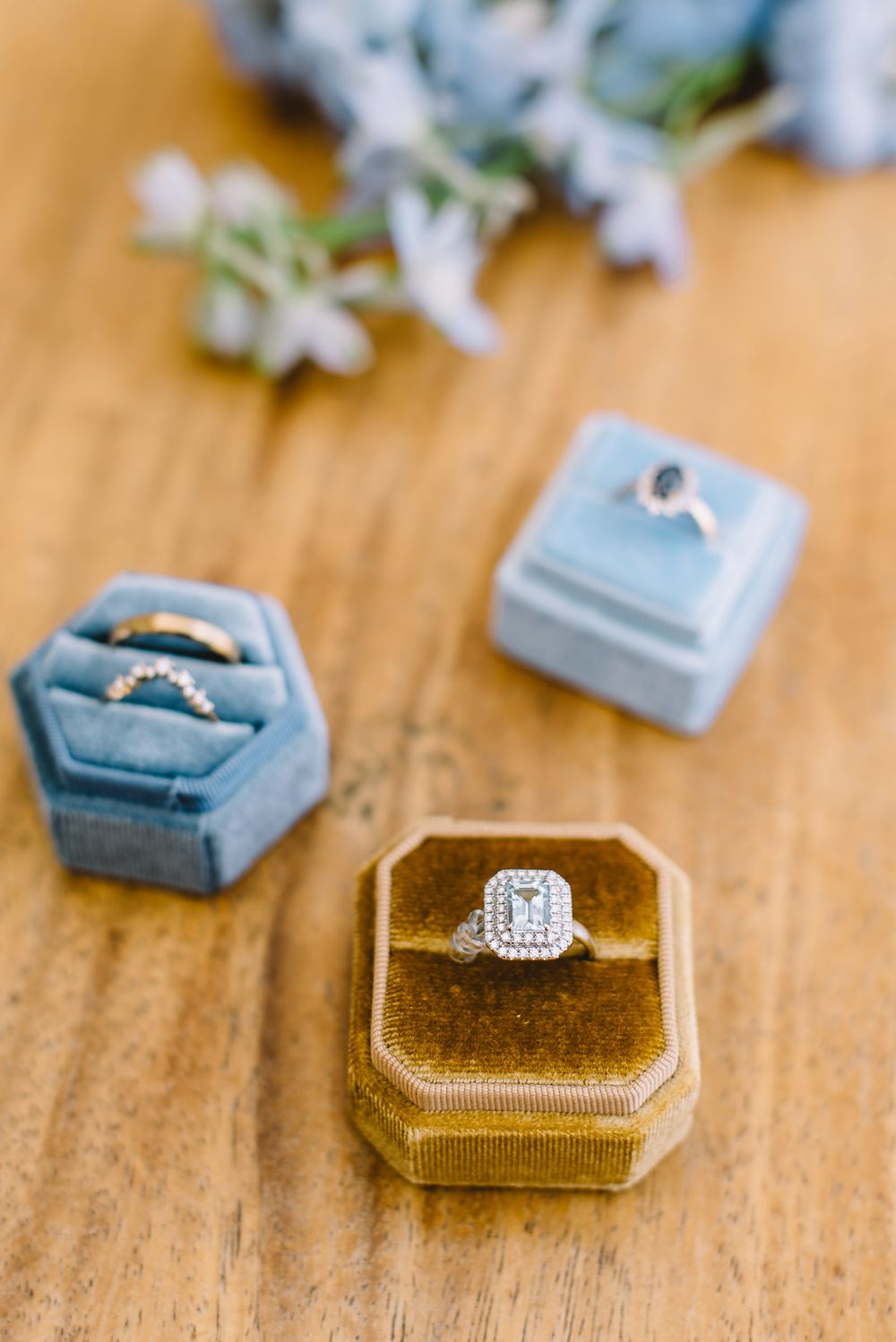 Three diamond rings displayed in velvet ring boxes on a wooden surface with soft floral accents.