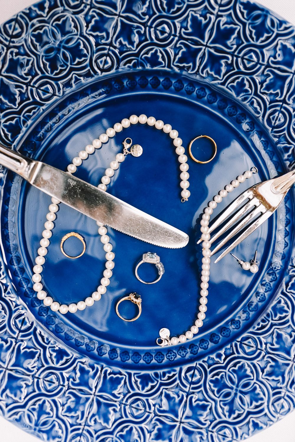 Blue ceramic plate with pearl necklace, gold rings, diamond ring, and silver cutlery arranged elegantly.