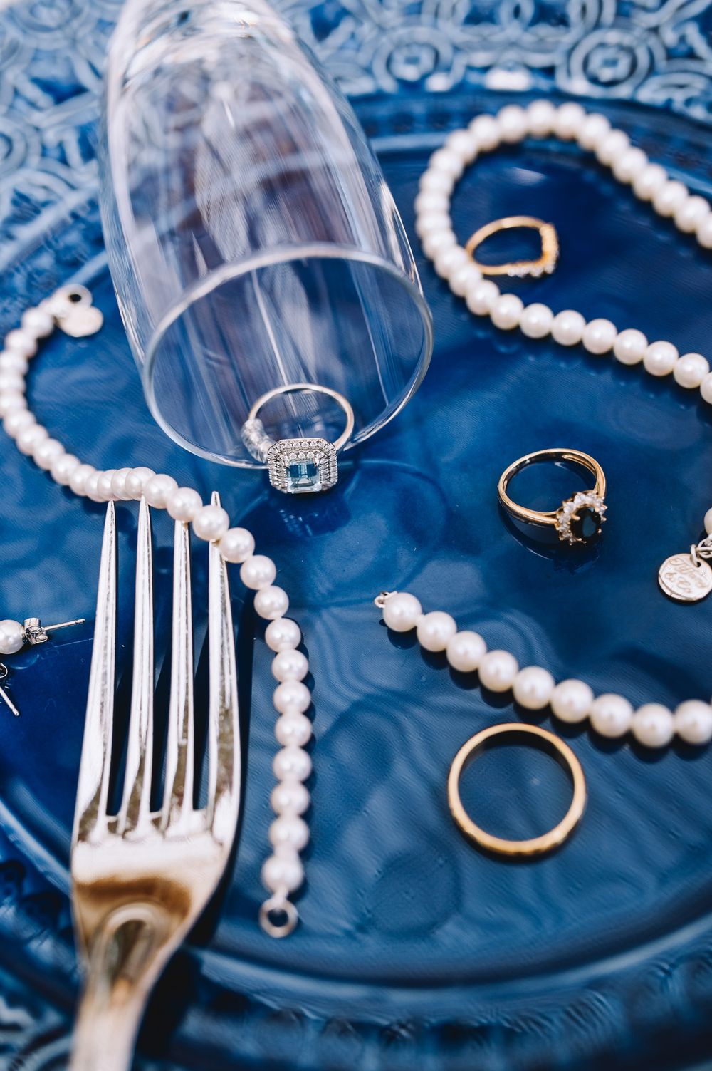 Gold rings and pearl necklaces arranged on a blue glass plate with a fork and champagne flute.