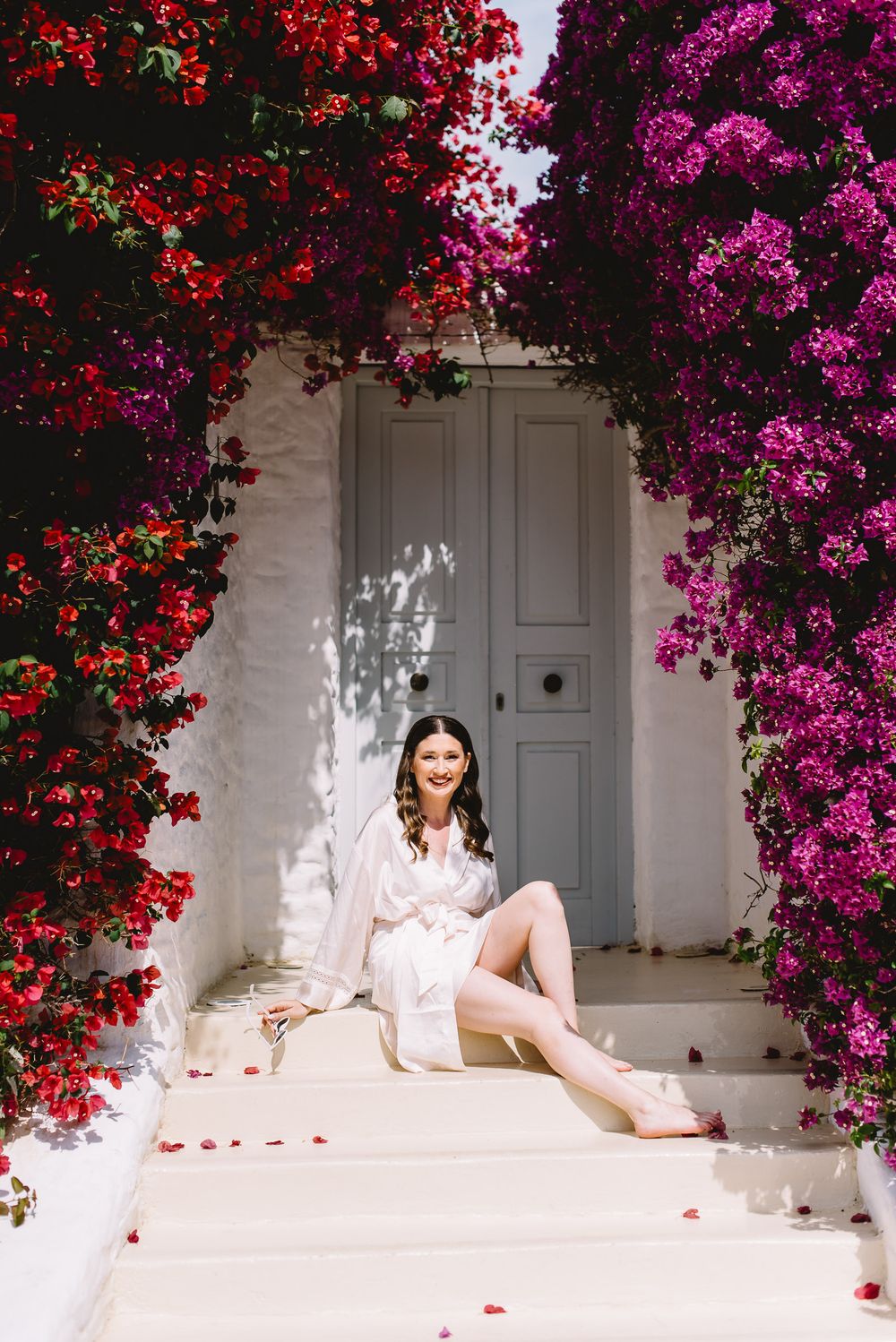 Bride in white silk robe sitting on steps surrounded by vibrant red and purple bougainvillea flowers.