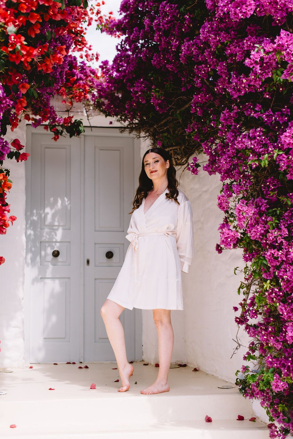 Woman in white dress standing barefoot under vibrant pink bougainvillea flowers by a grey door.
