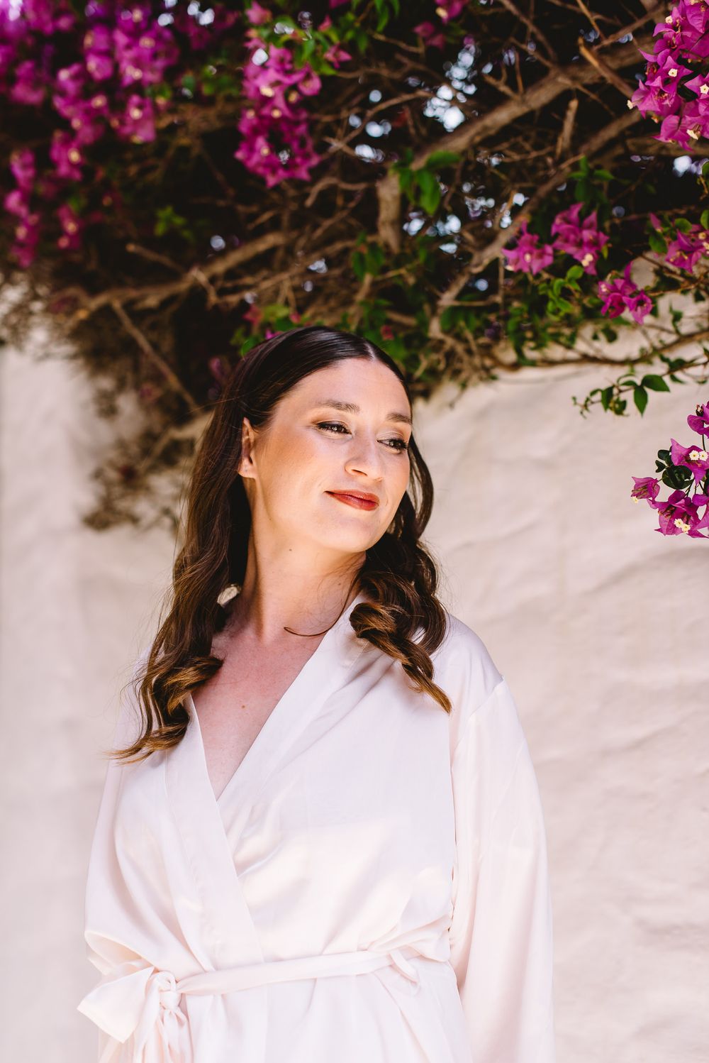 Bride in white robe gazing up at pink bougainvillea blooms against a white wall.