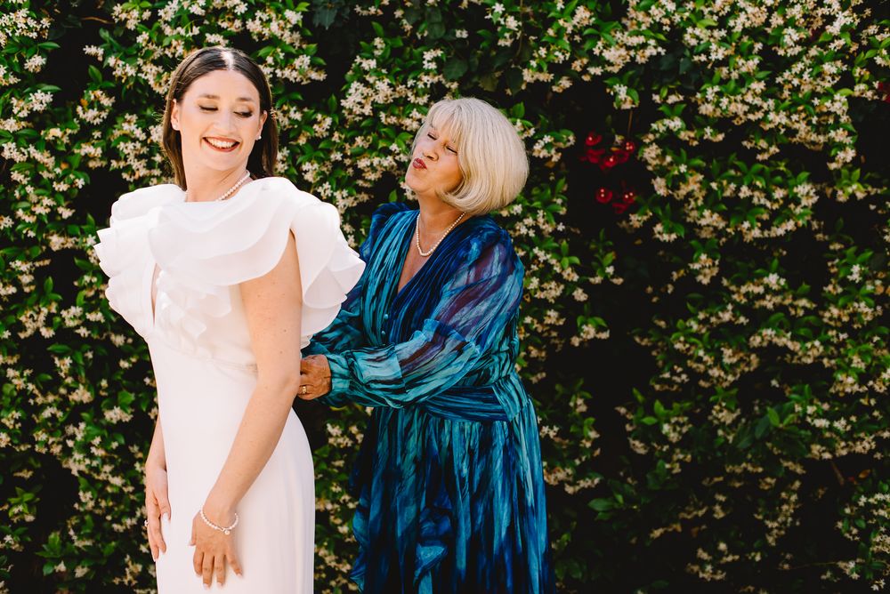 Bride in white gown with older woman in teal dress smiling together in front of a lush floral green wall.