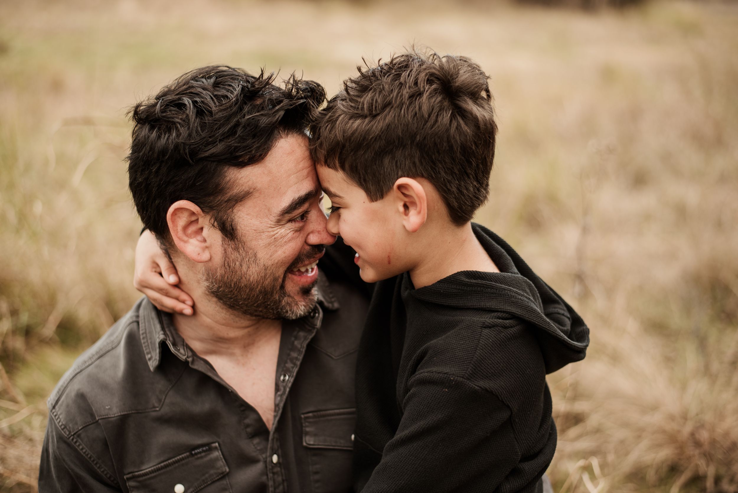 A father and young son share a tender forehead-to-forehead moment, smiling outdoors in a grassy field.