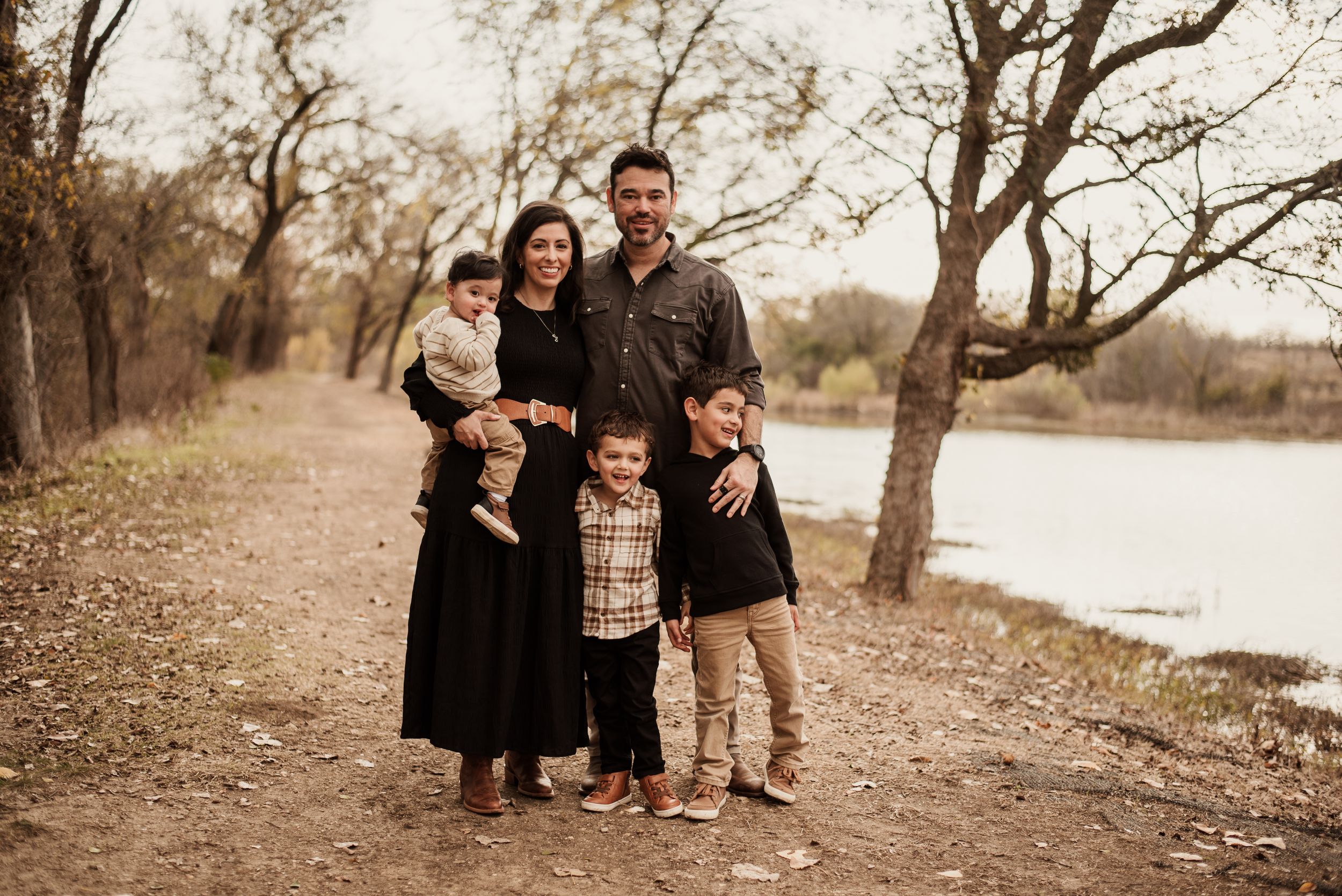 Family of five posing outdoors near a river surrounded by bare trees in autumn.