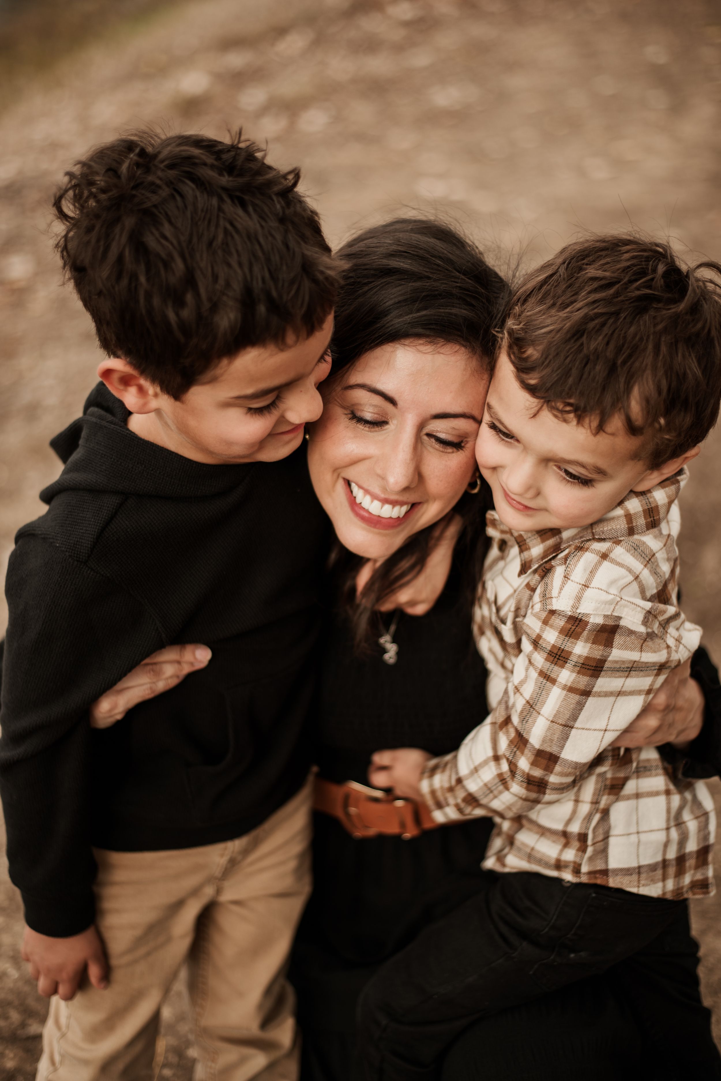 A smiling woman hugs two young boys in a warm outdoor family portrait with autumn tones.