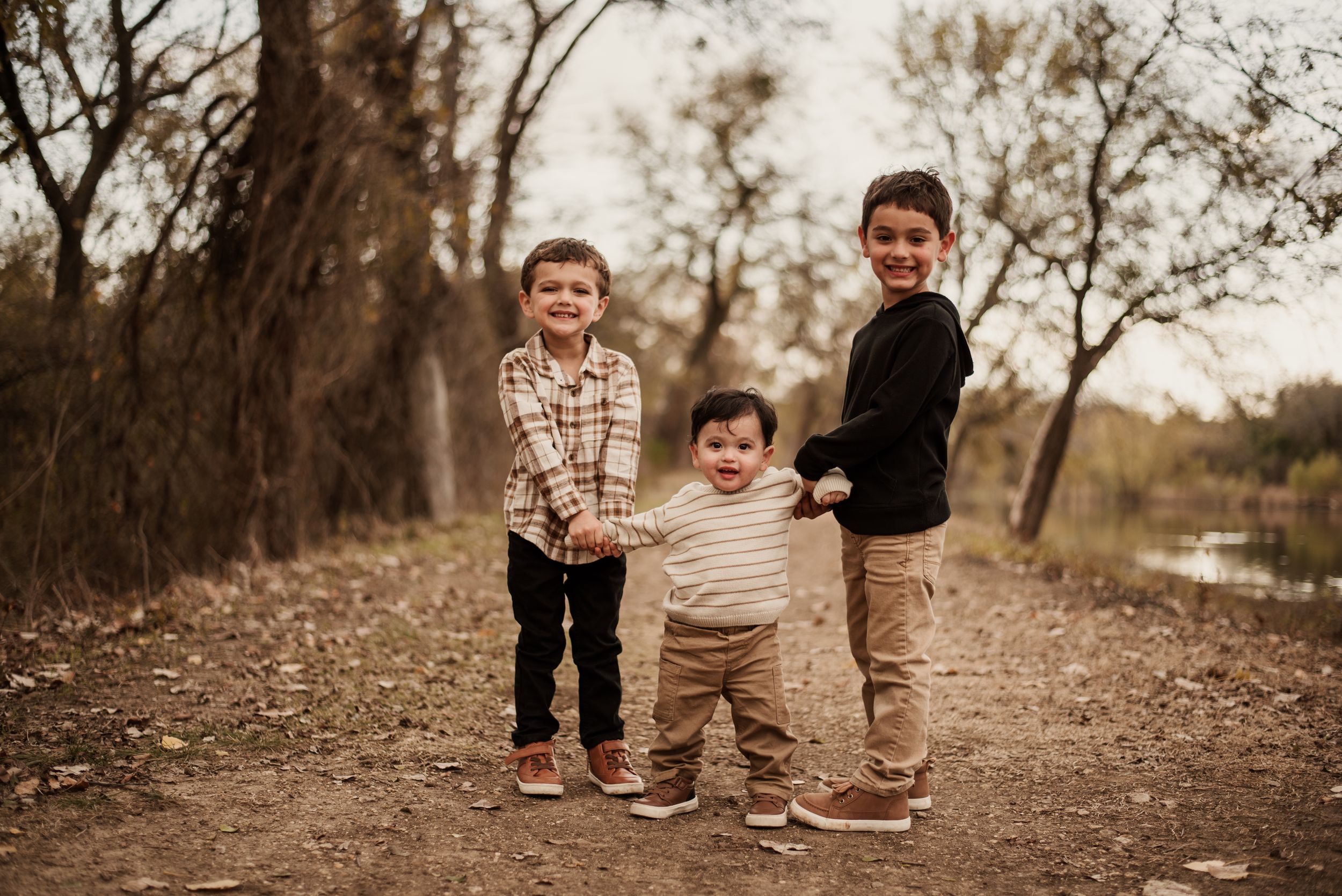 Three young children smile and hold hands on a dirt path surrounded by autumn trees in a warm outdoor family portrait.