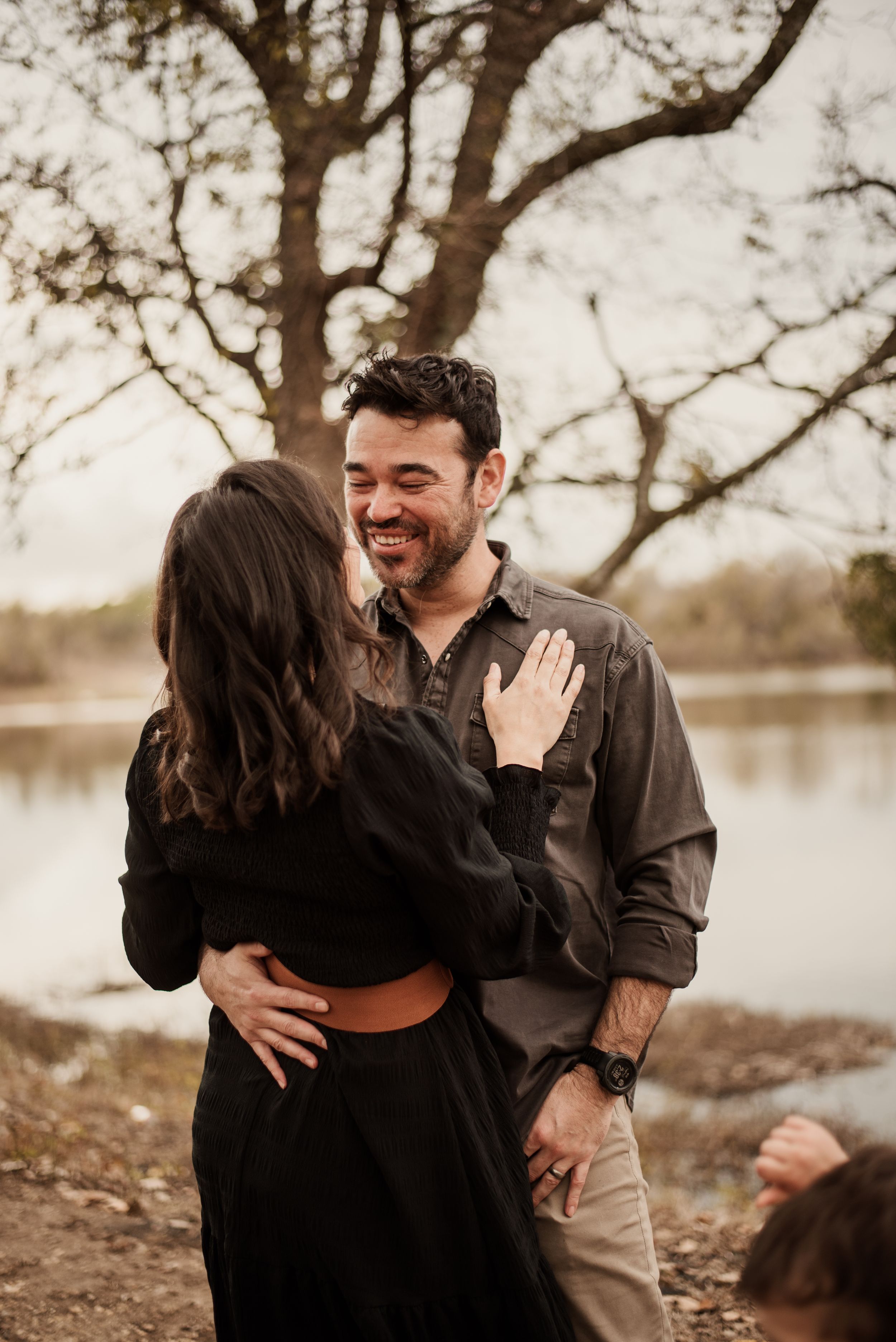 A smiling bearded man embraces a dark-haired woman outdoors near a lake, surrounded by bare trees in autumn.