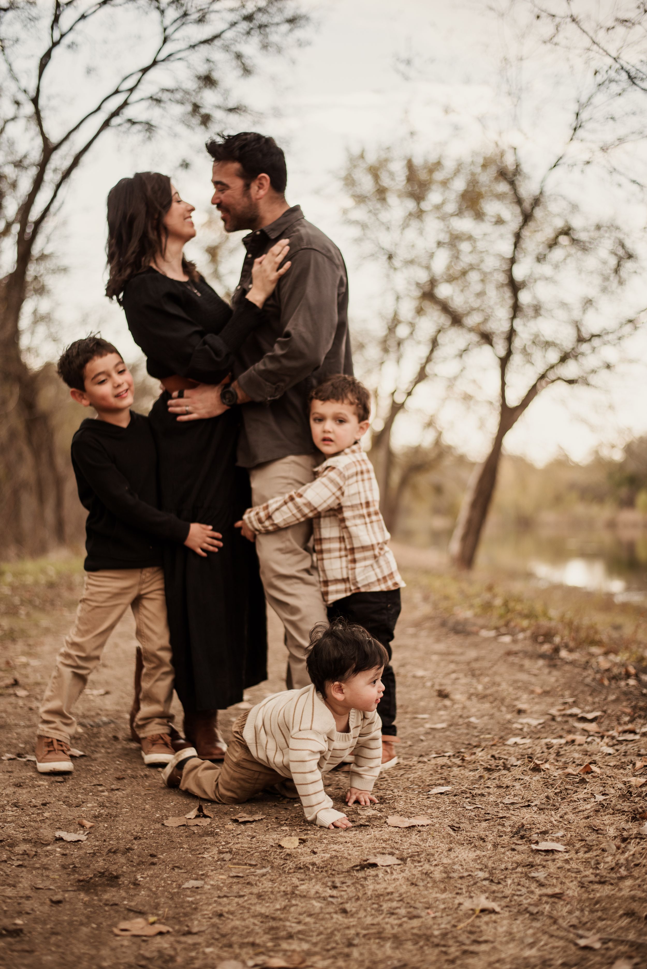 Family of five posing on a dirt path surrounded by bare autumn trees, parents embracing while three young boys gather around them.