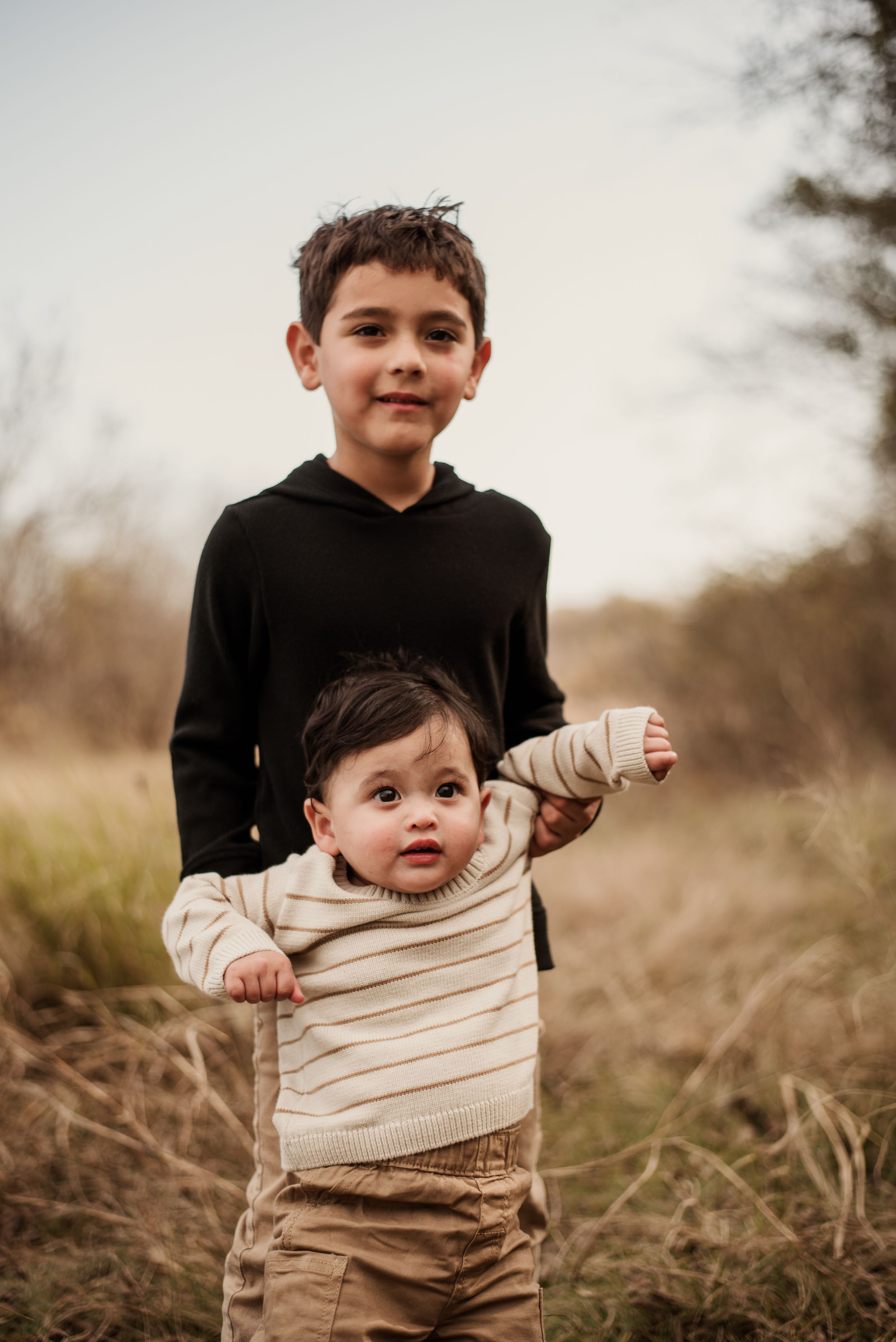 Older boy in black hoodie helping baby sibling stand in an outdoor field during a family photo session.