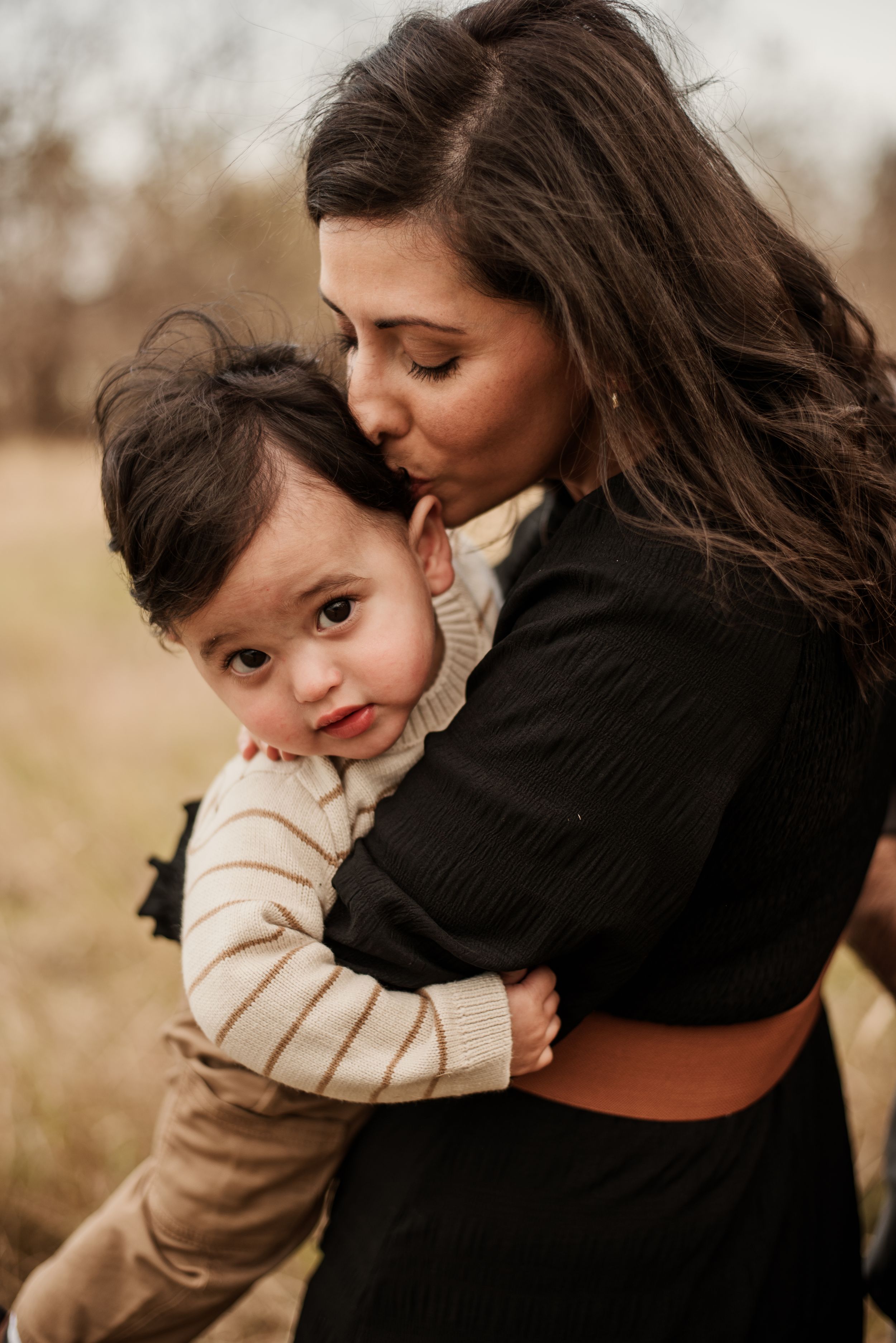 A mother tenderly kisses her toddler while holding them close outdoors in a field.