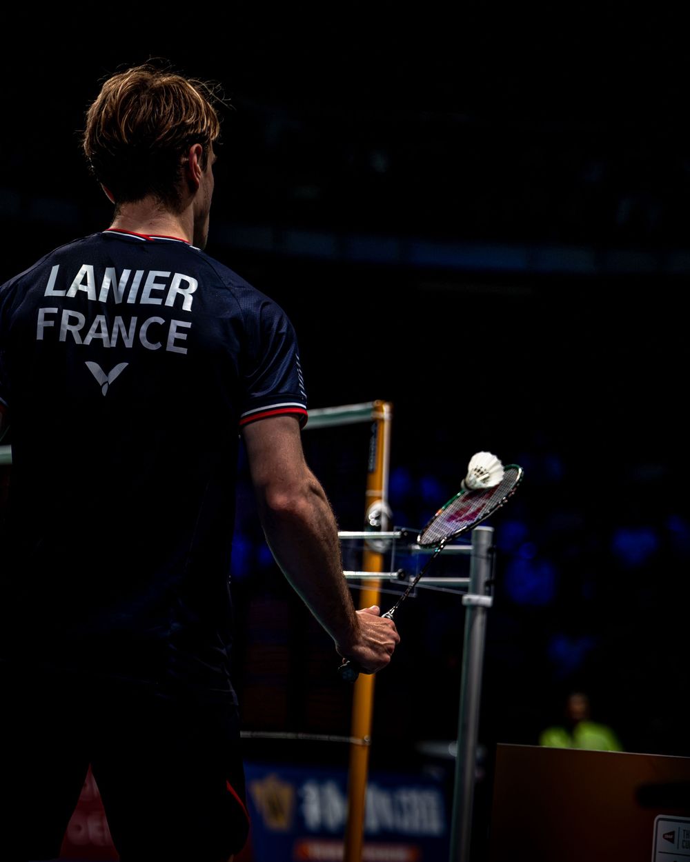French badminton player Lanier seen from behind holding a racket during a competitive match wearing France team jersey.