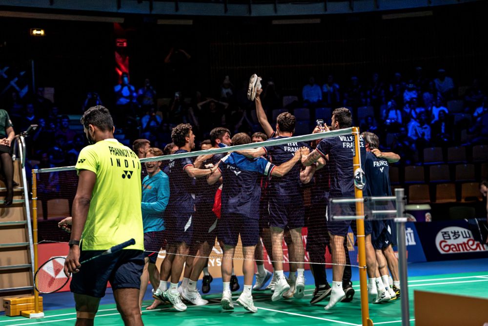Badminton team celebrating victory on court, players huddle near net in arena with spectators watching.