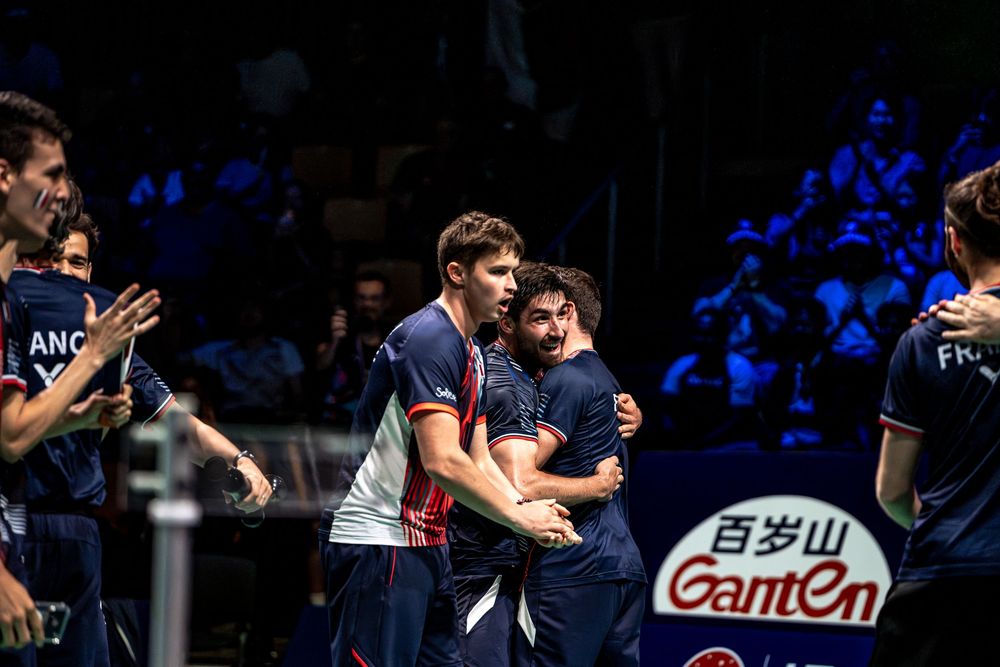 Badminton players in navy uniforms celebrate together on court at an international tournament with Ganten sponsorship banners.