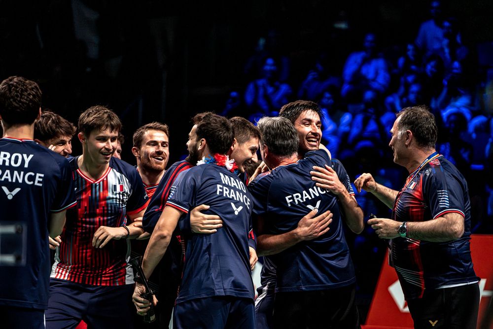 French badminton team celebrating victory together on court, wearing navy blue France jerseys.