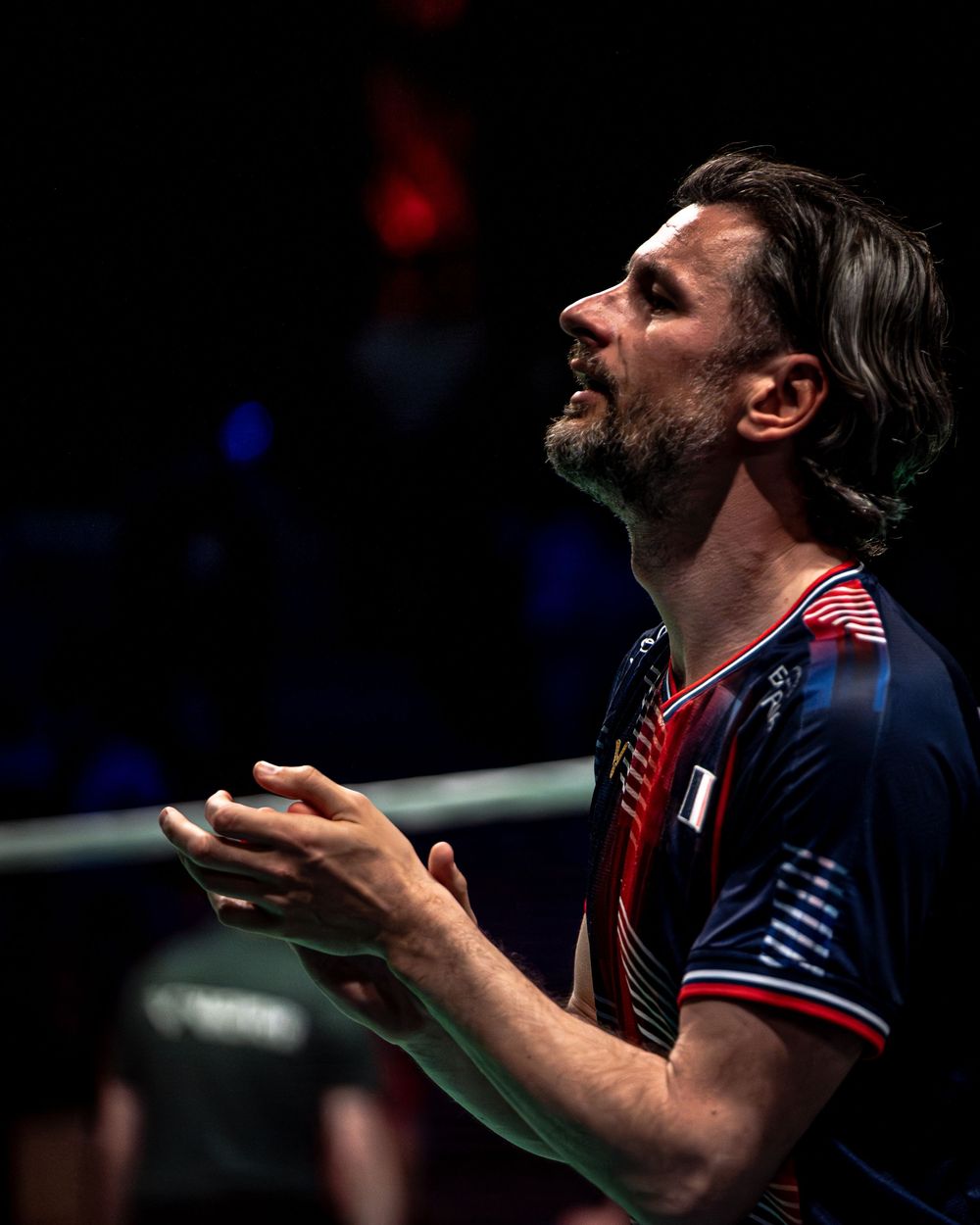 Bearded male badminton player in navy uniform reacting emotionally during a competitive match indoors.