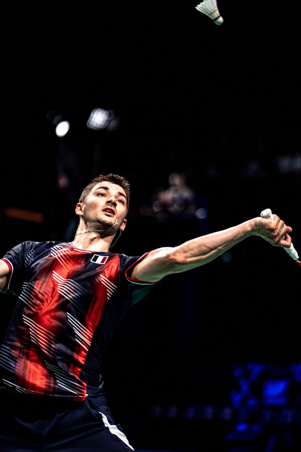 Badminton player in red and navy kit stretches to hit a shuttlecock during a competitive match.