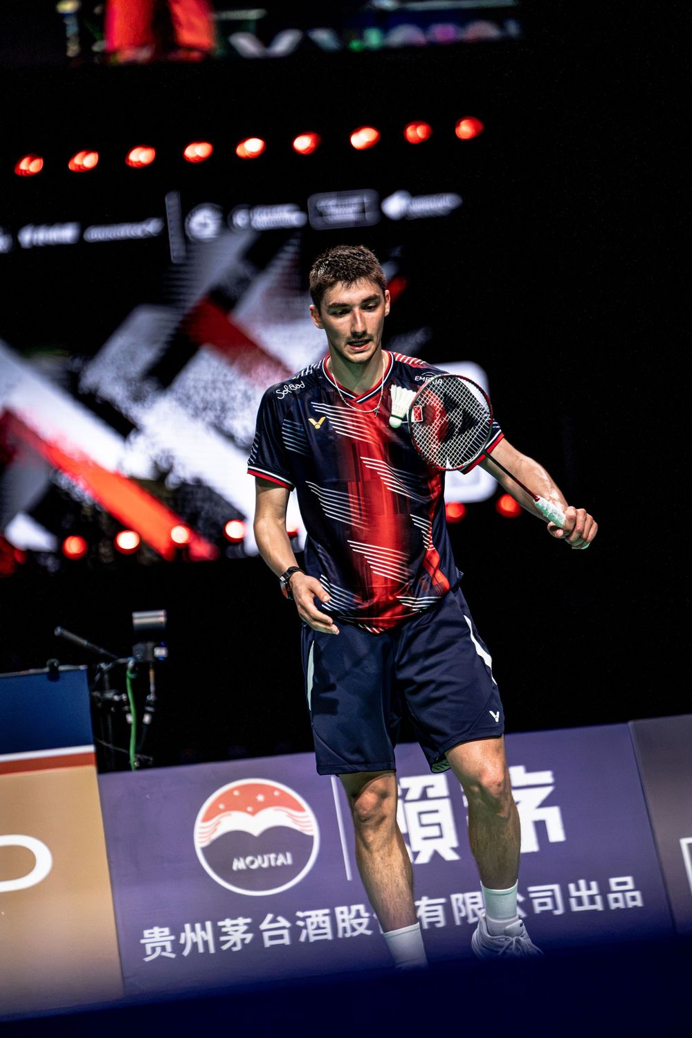 Badminton player in navy and red Victor kit holds racket on court at professional tournament with Chinese sponsors.