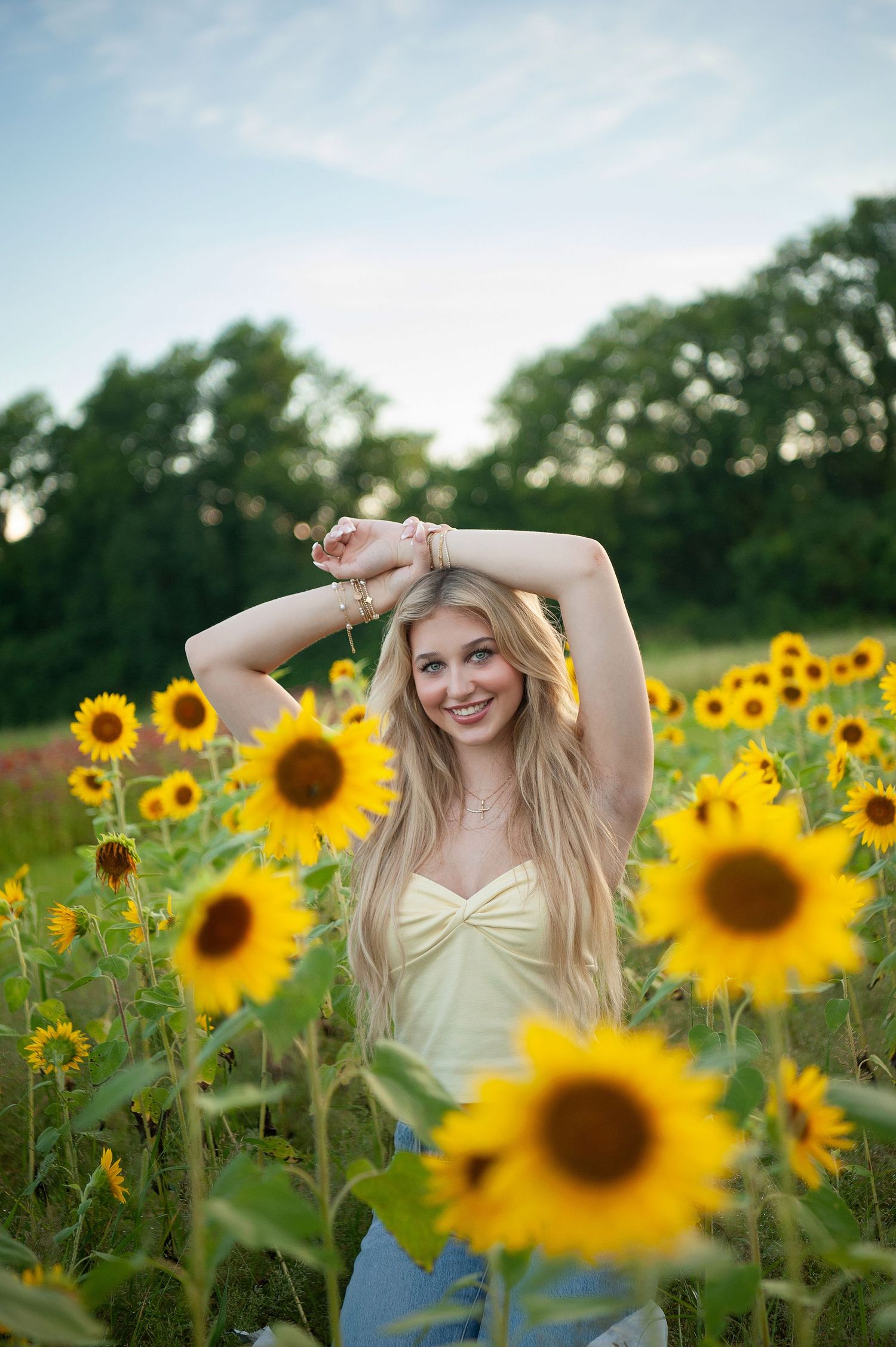 Smiling blonde woman in yellow top posing gracefully in a vibrant sunflower field with trees in the background.