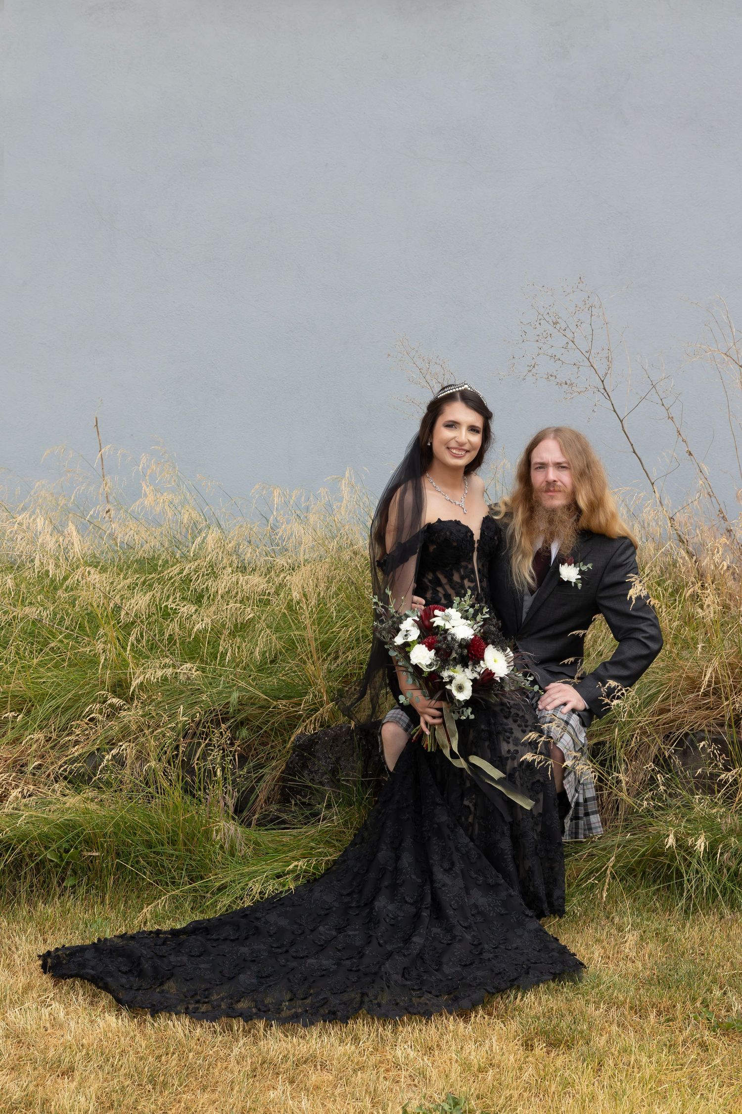 bride wearing black sitting on groom's lap in front of gray wall looking at the camera smiling Milestones Studios LLC | Milestones Portraits