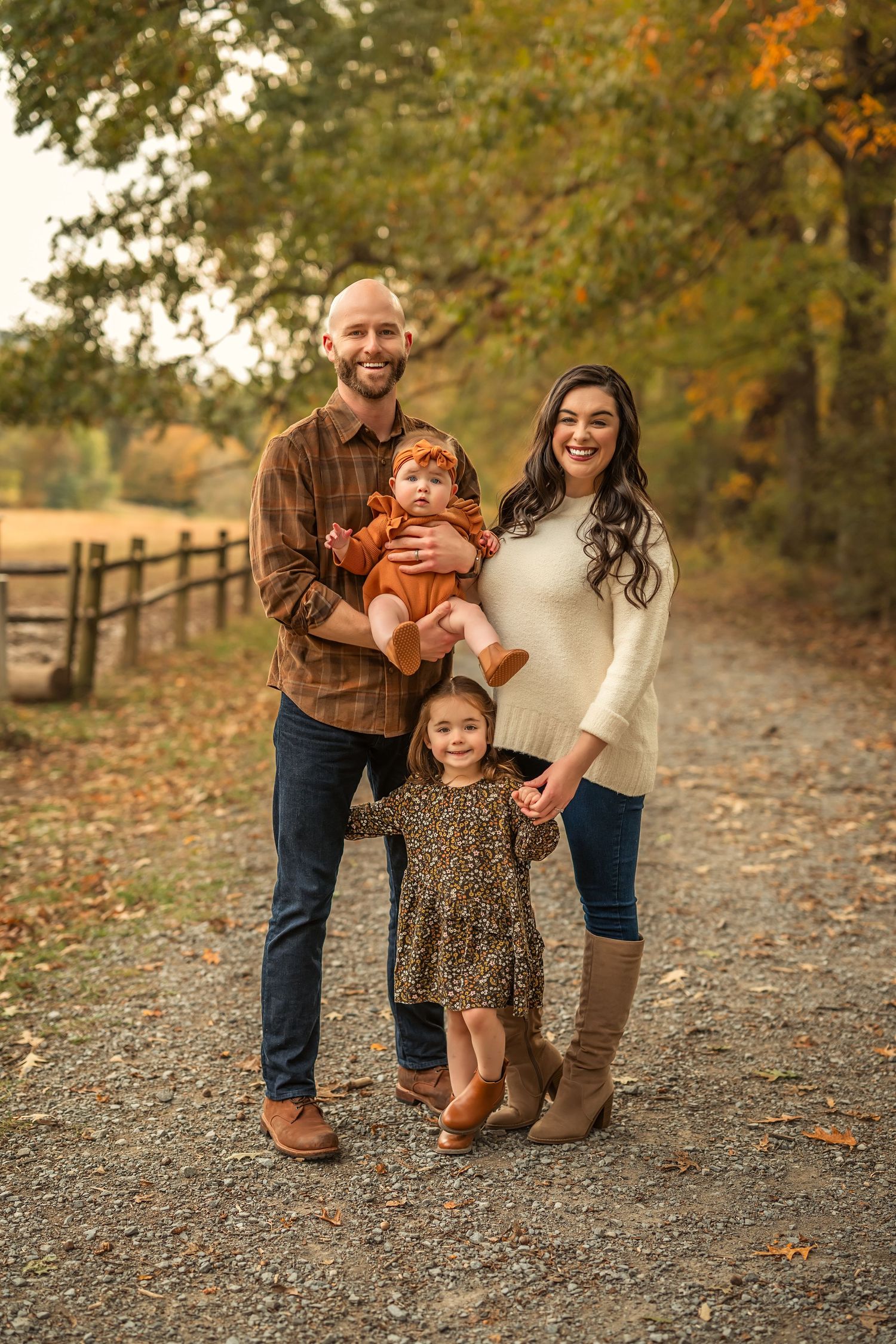 Family of four posing on a tree-lined path in autumn, parents holding young children in warm fall-colored outfits.