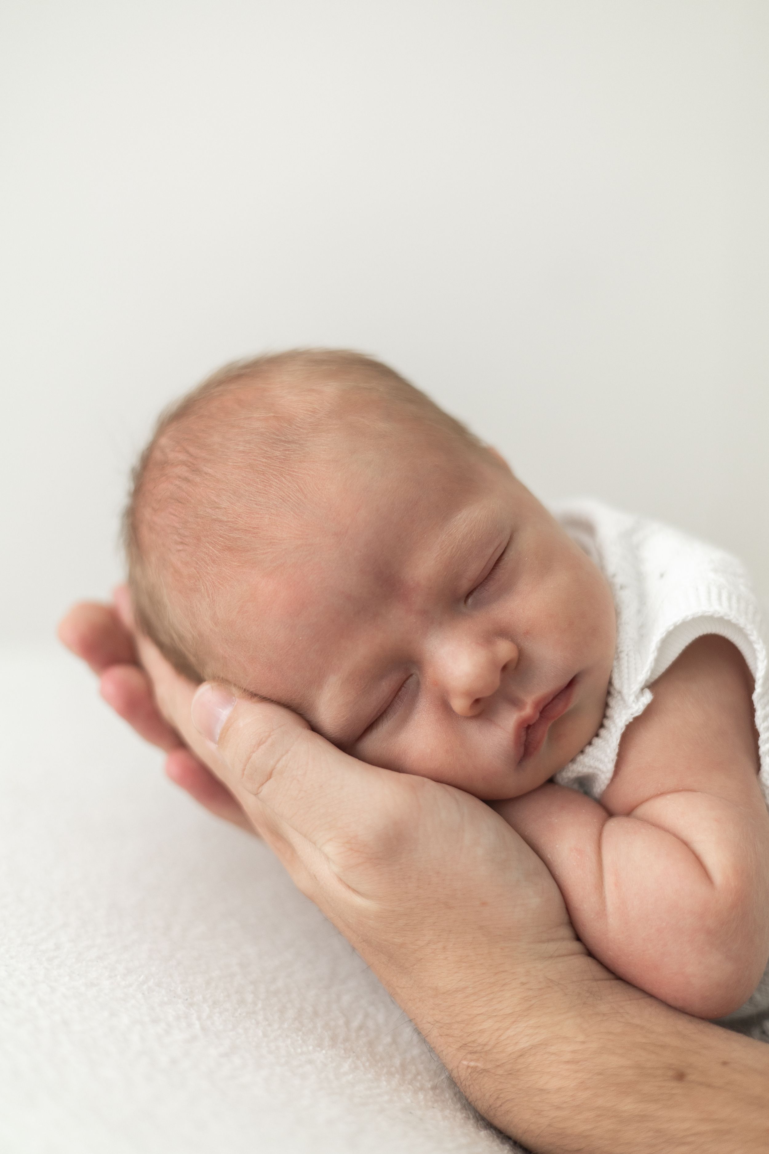 Sleeping newborn baby cradled gently in Dads hands, wearing white outfit on soft white background natural newborn photography Nottingham
