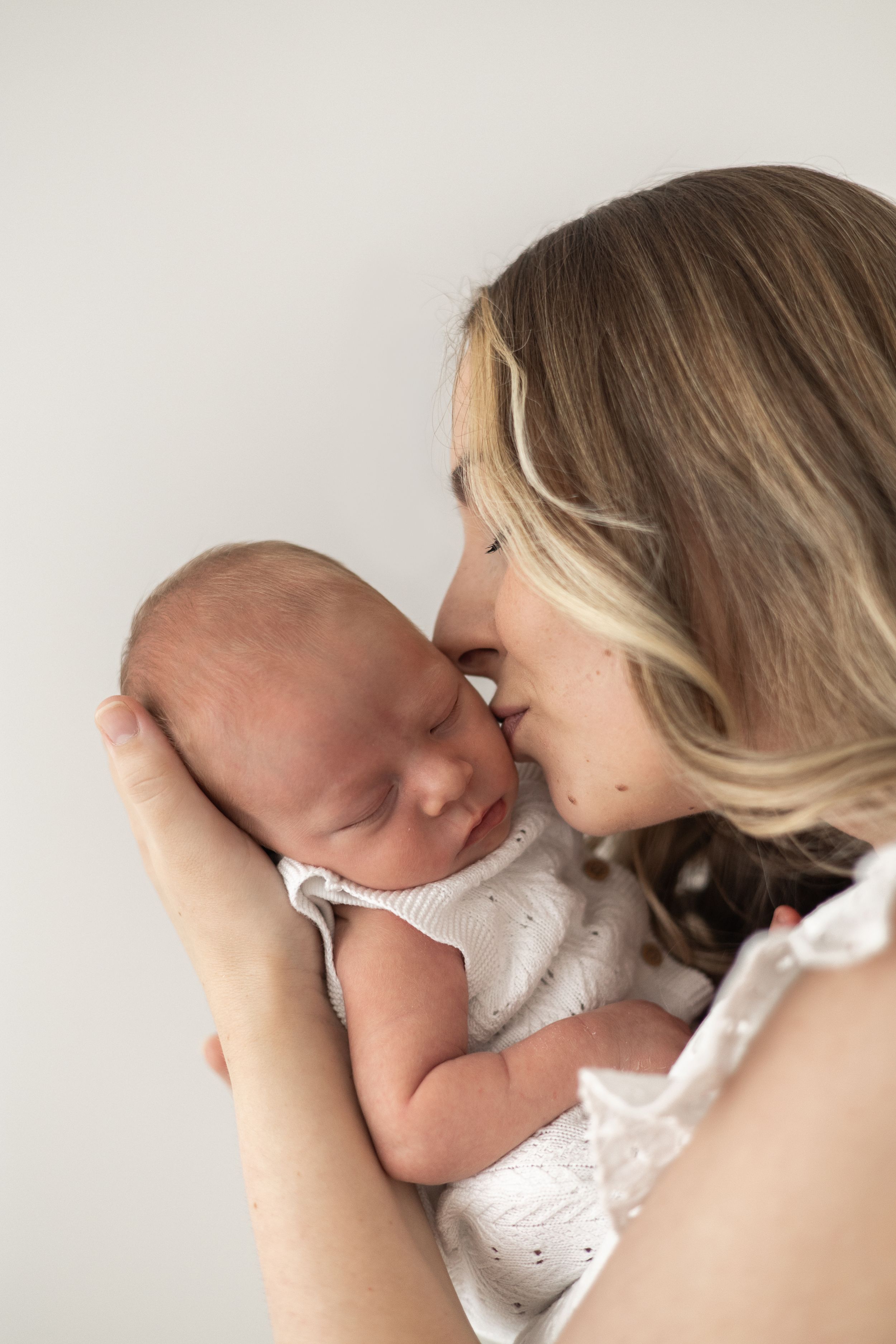 A loving mother gently kisses her sleeping newborn baby, against a soft white background natural newborn photography Nottingham