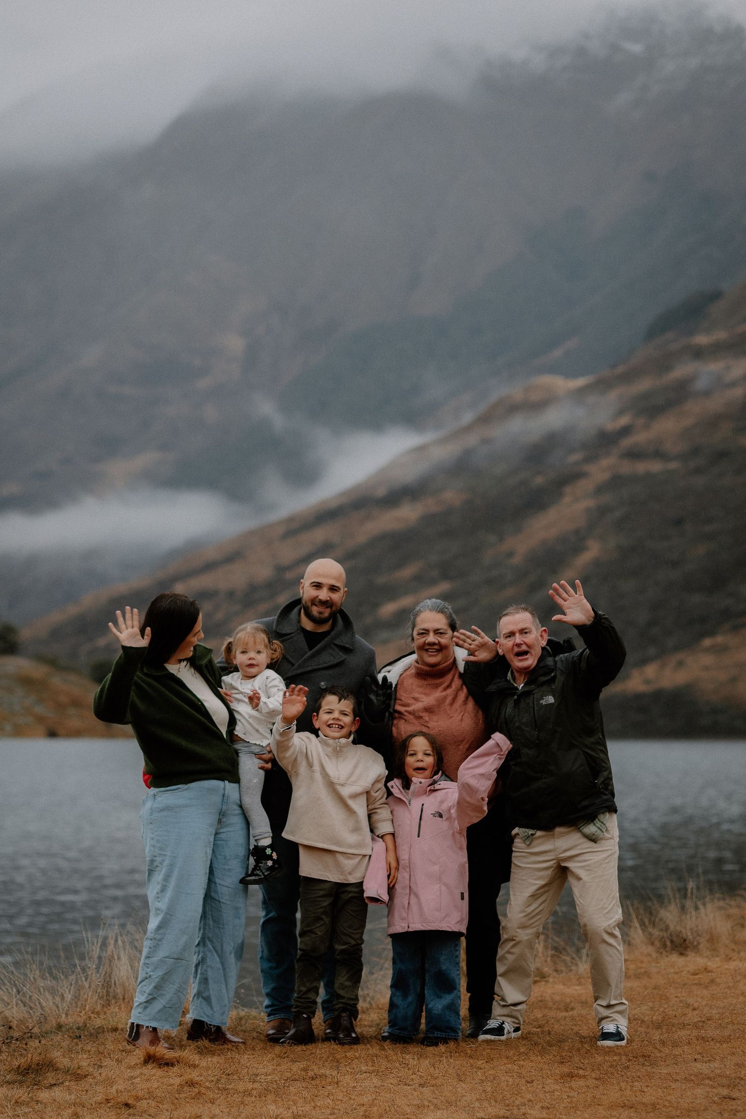 Multi-generational family waving and smiling by a mountain lake on a cloudy day in a scenic outdoor setting.