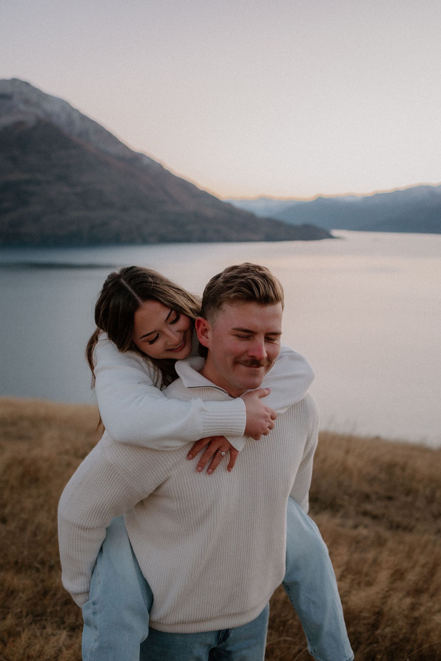 Couple sharing a piggyback ride at golden hour near a mountain lake, both wearing white sweaters.
