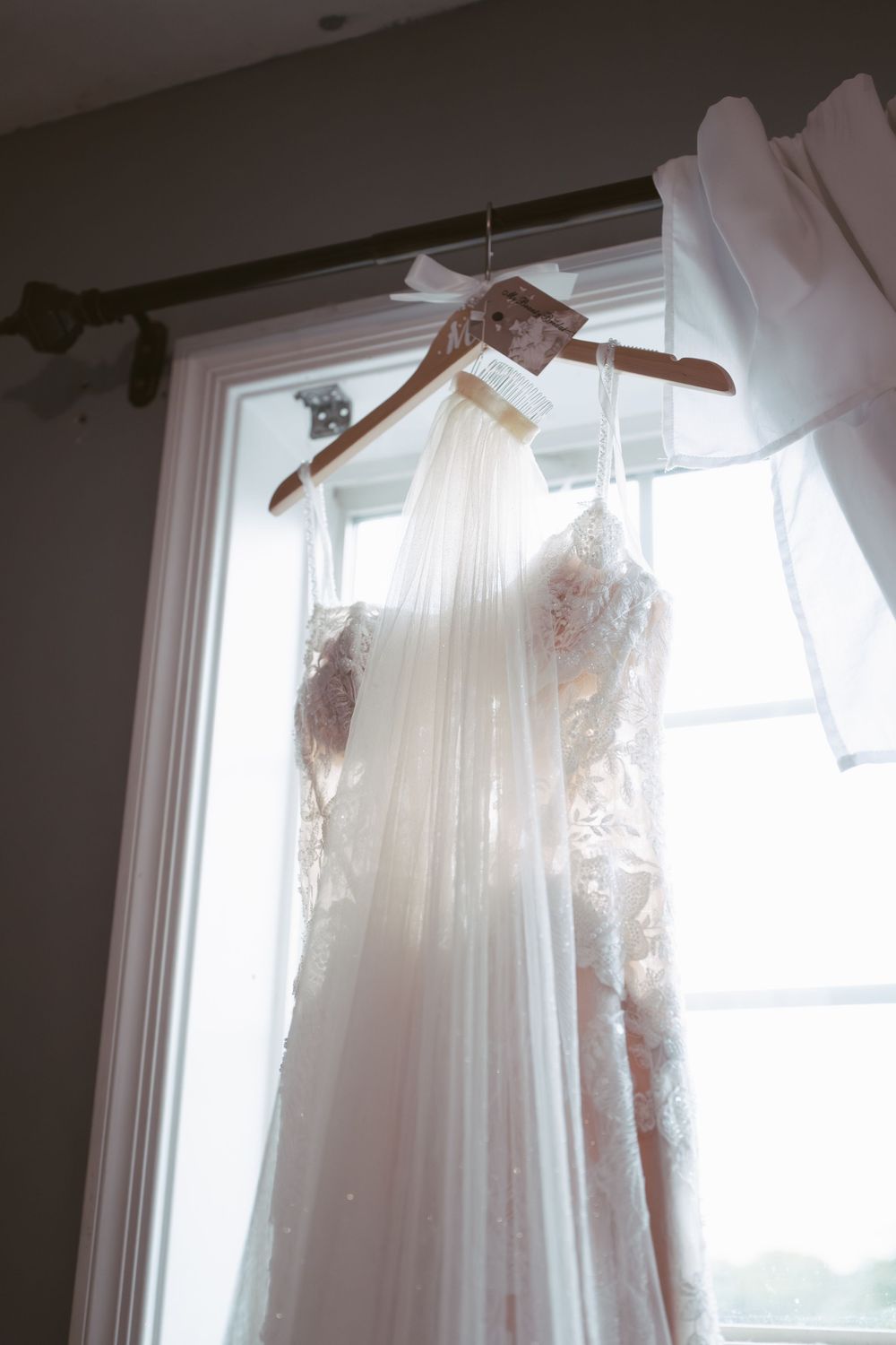 Lace wedding dress with veil hanging on wooden hanger in front of bright window.