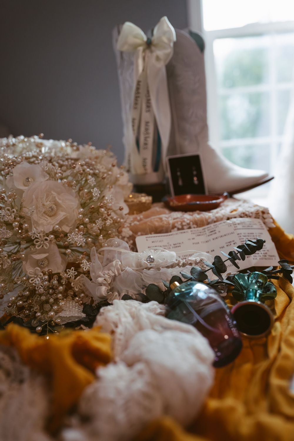Bridal flat lay with white cowboy boots, floral bouquet, veil, jewelry, and perfume on golden fabric.